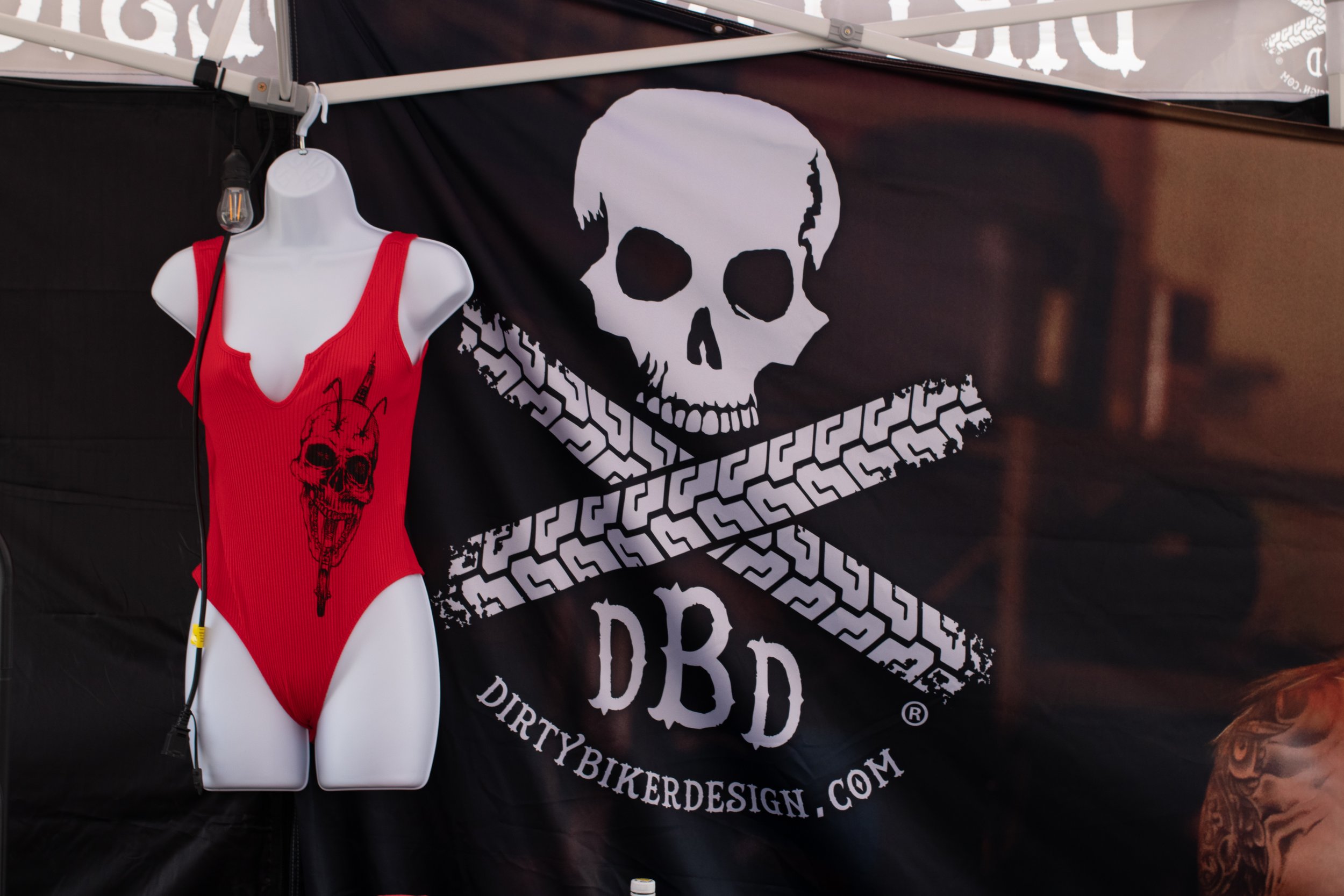 A black flag with a white skull and crossbones design and the text 'DIRTY BIKER DESIGN' in white. A mannequin torso wearing a red bodysuit with a black skull design is in front of the flag, with a wire connected to the bodysuit's shoulder. Part of a 
