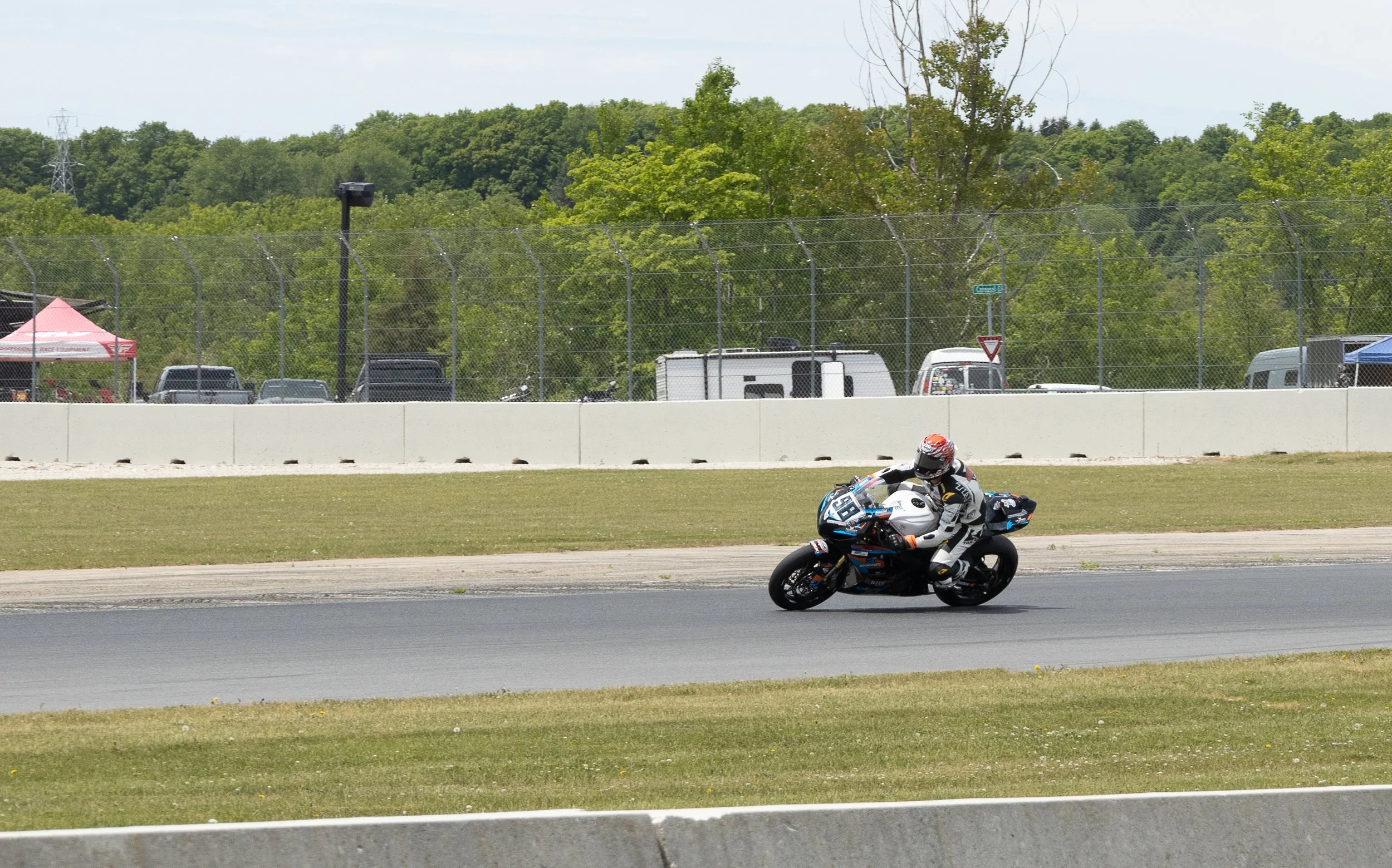 Motorcycle racer in full gear leaning into a turn on a racetrack, with a background of green trees, vehicles parked behind a fence, and a tent.