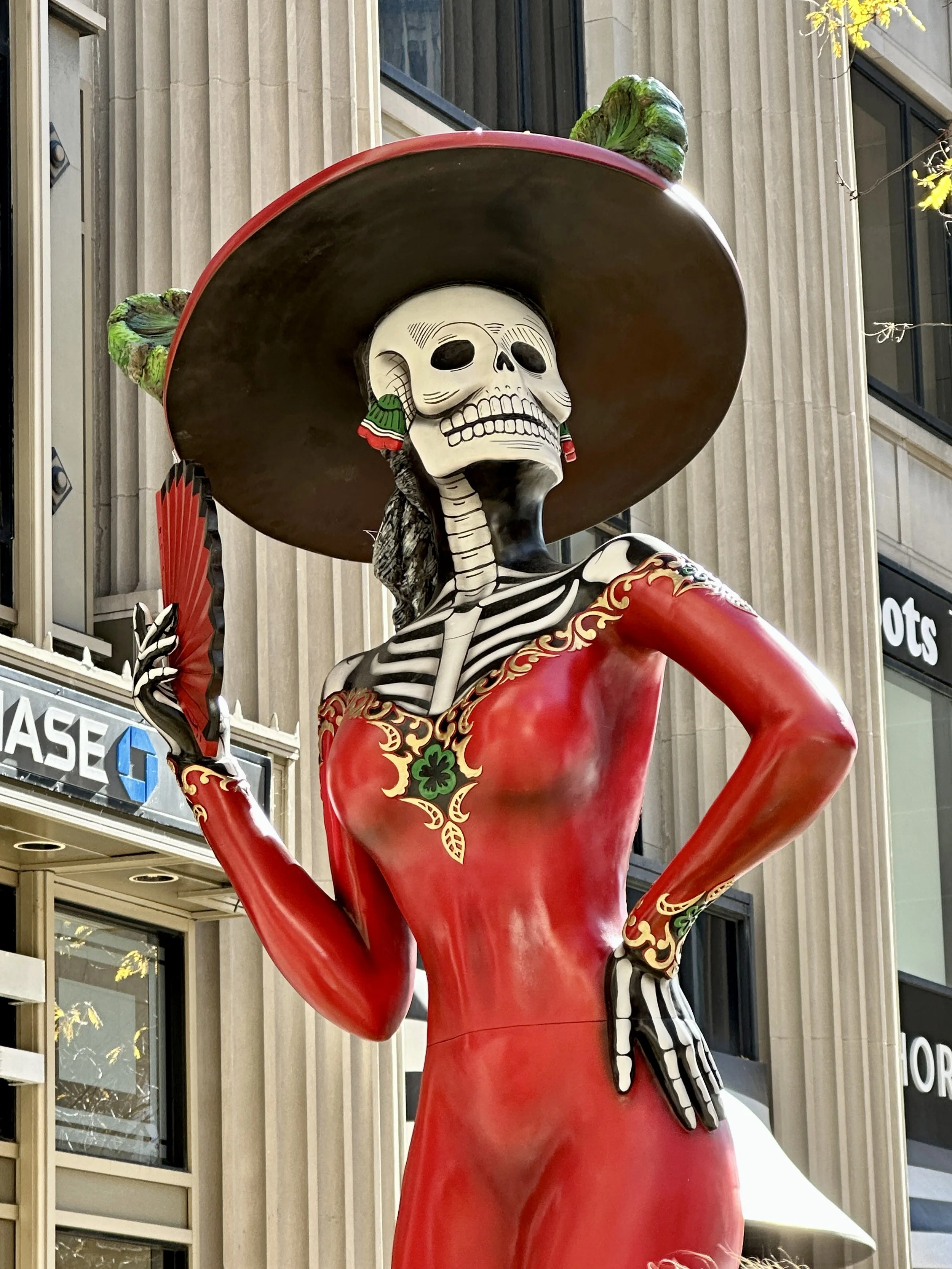Statue of a woman with a large sombrero, skeleton makeup, and dressed in a red dress with gold details, standing against a city building background.