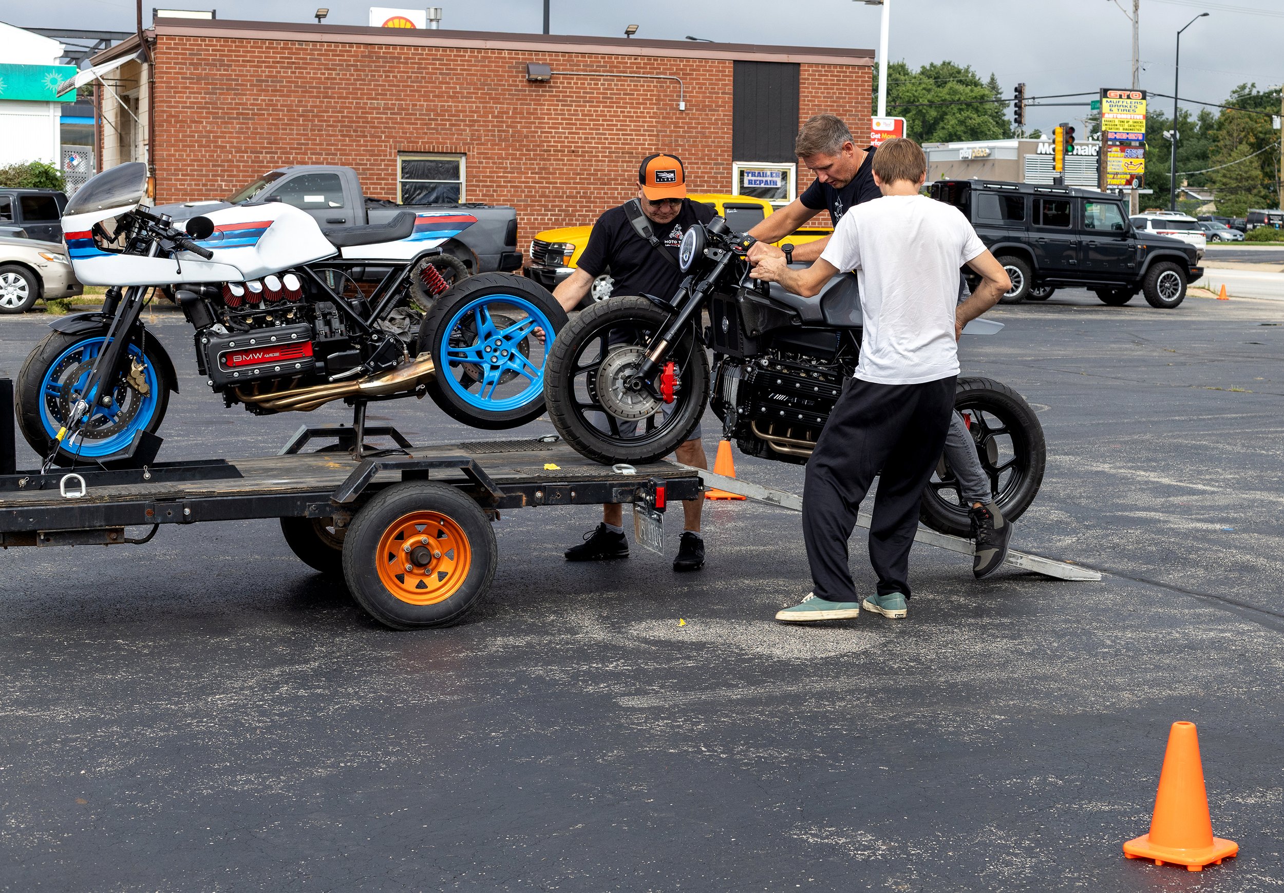 Three men working on a black motorcycle on a trailer in a parking lot, with another colorful motorcycle beside them and orange cones nearby.