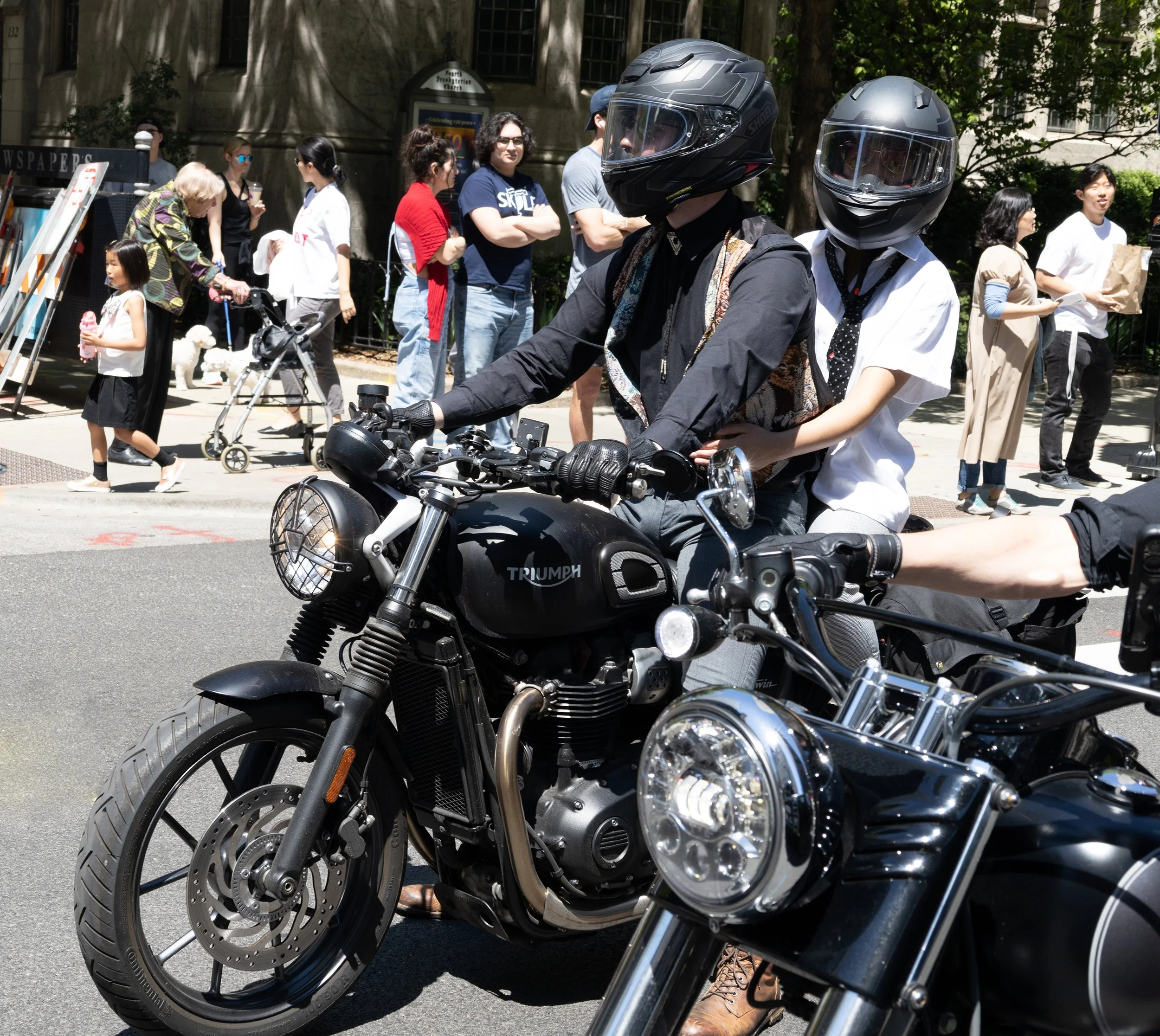 People riding motorcycles in a busy city street with pedestrians and buildings in the background.