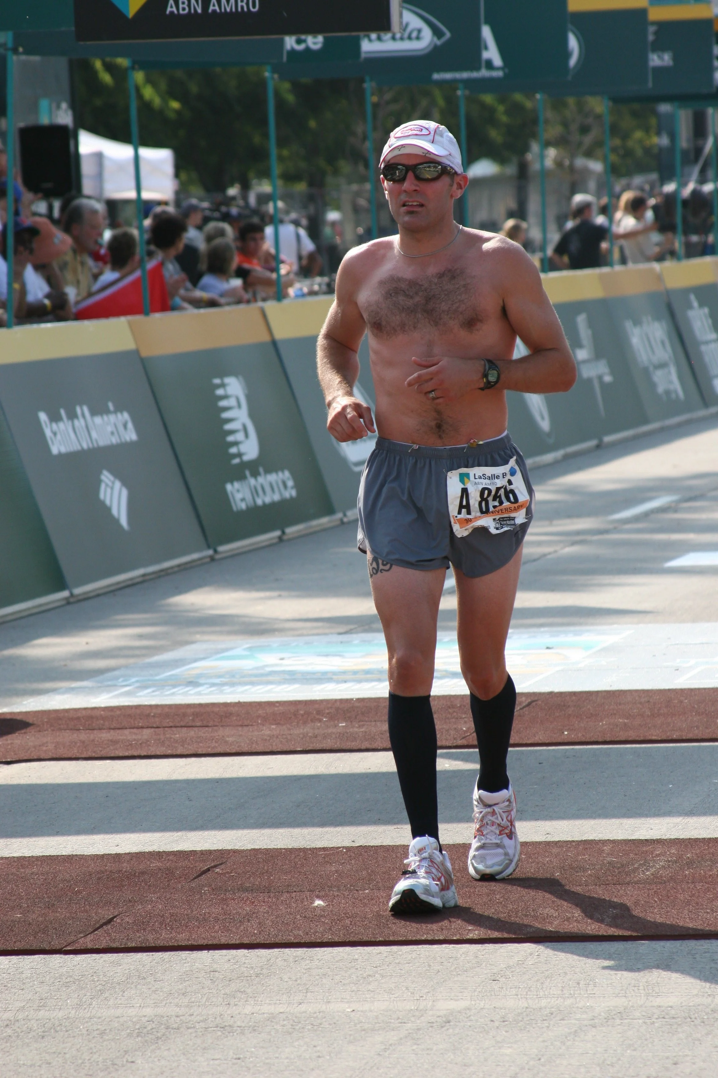 A man running in a marathon, shirtless, wearing shorts, sunglasses, a white cap, black socks, and white running shoes, on a track with spectators behind barriers and banners overhead.
