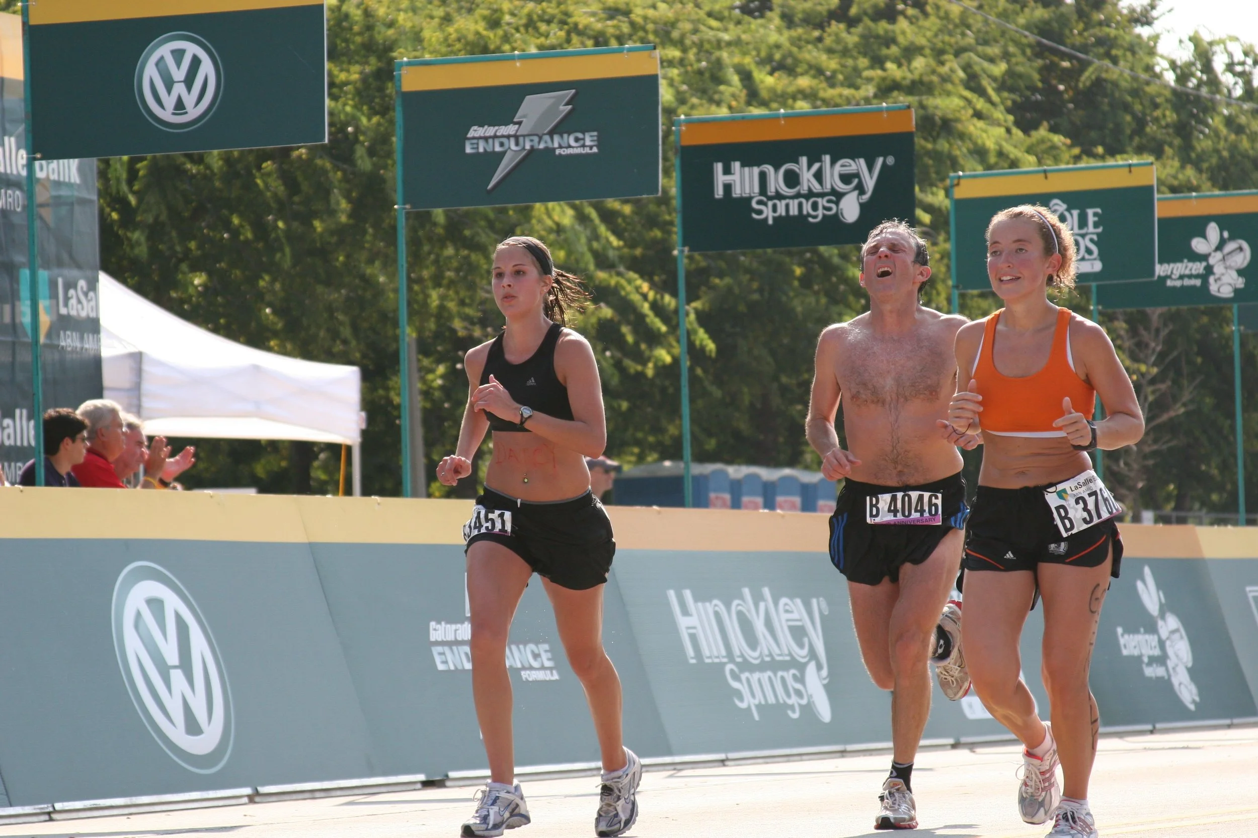 Three runners, two women and a man, participating in a race near the finish line with banners and spectators in the background on a sunny day.
