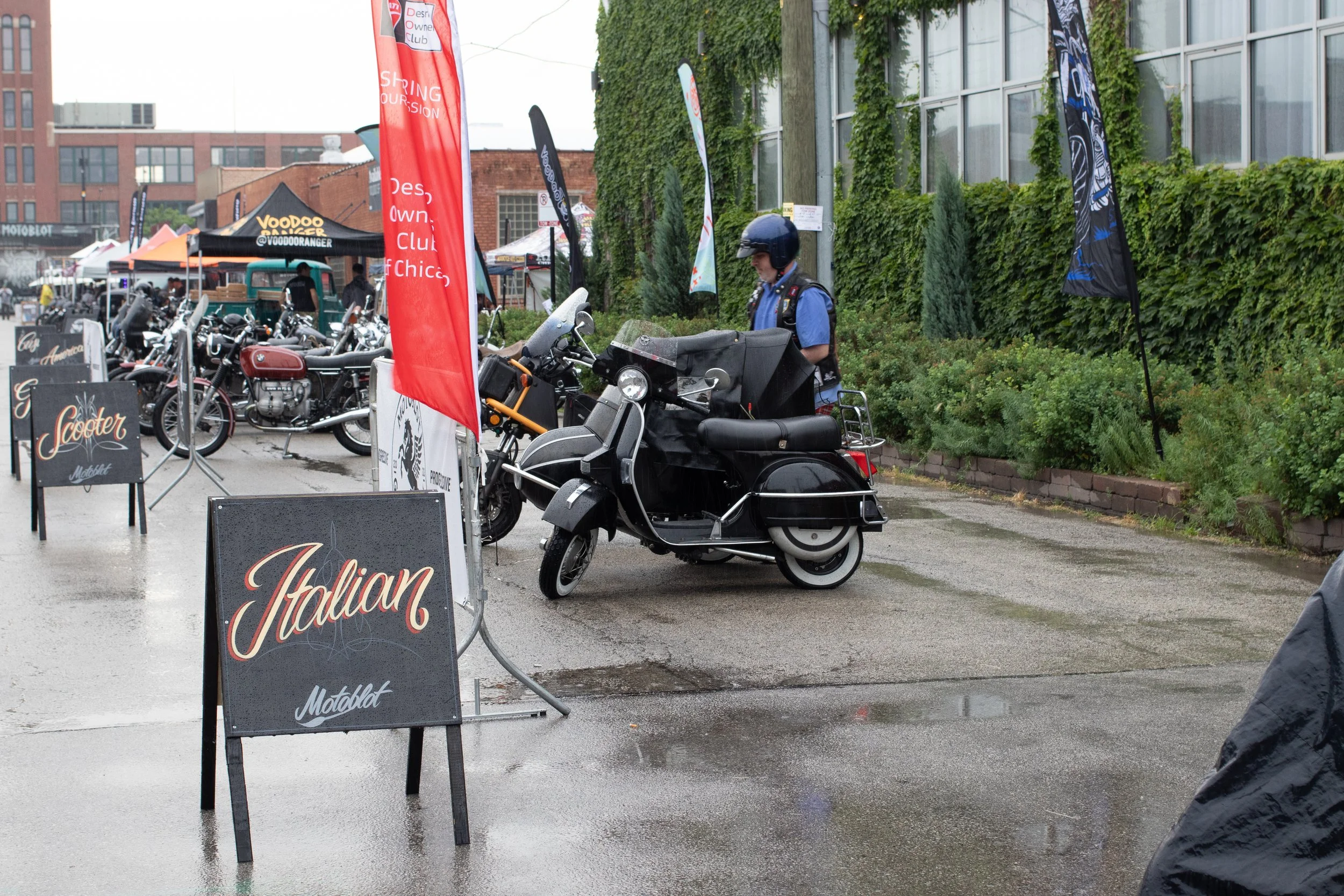 A street scene with a motorcycle and scooter parked in front of a sidewalk cafe or event area with banners and signs. A person in a blue uniform and helmet stands near the motorcycle. The background shows buildings with green ivy growing on a wall.