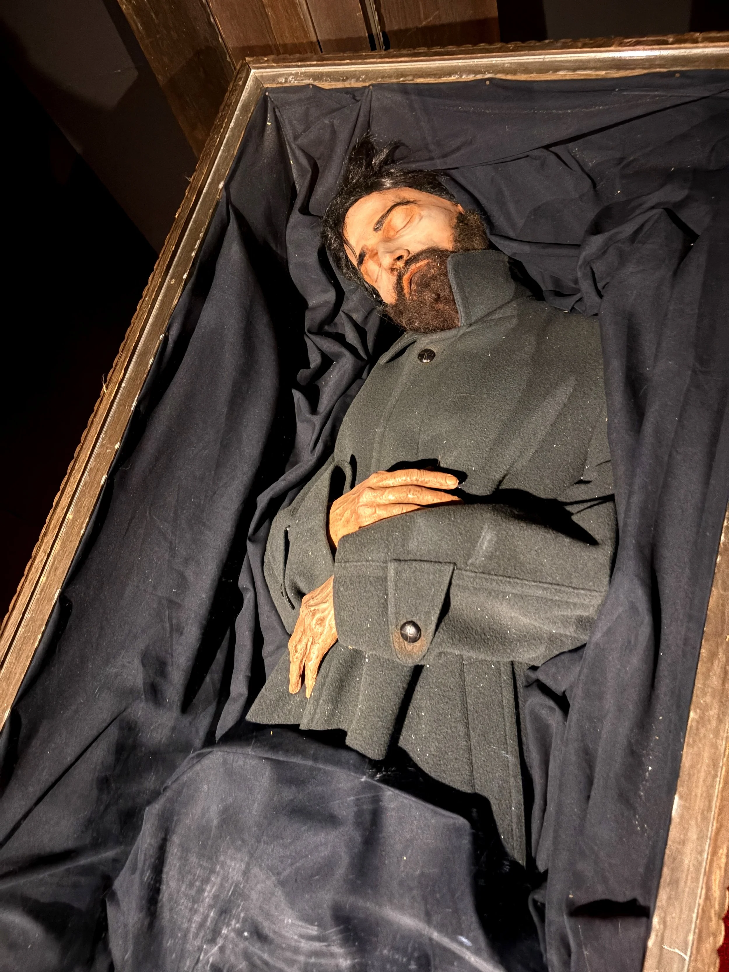 A wax figure of a man with long dark hair, a beard, and closed eyes lying in a coffin lined with black fabric and wooden edges.