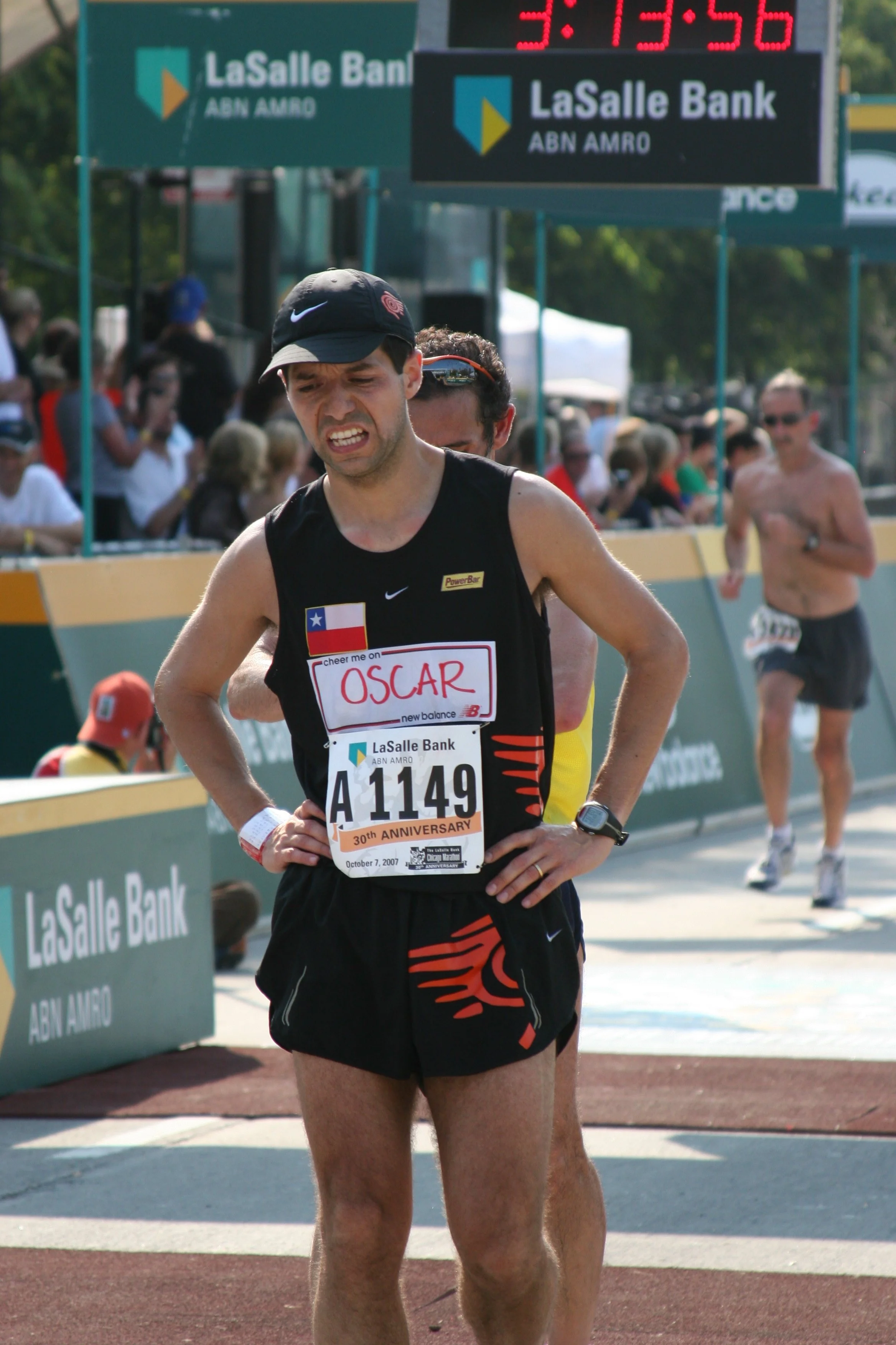 A male marathon runner wearing a black cap and a black singlet with a Texas flag and a bib number A1149, standing during a race with a pained expression. There are spectators and other runners in the background, with signs and a digital clock showing