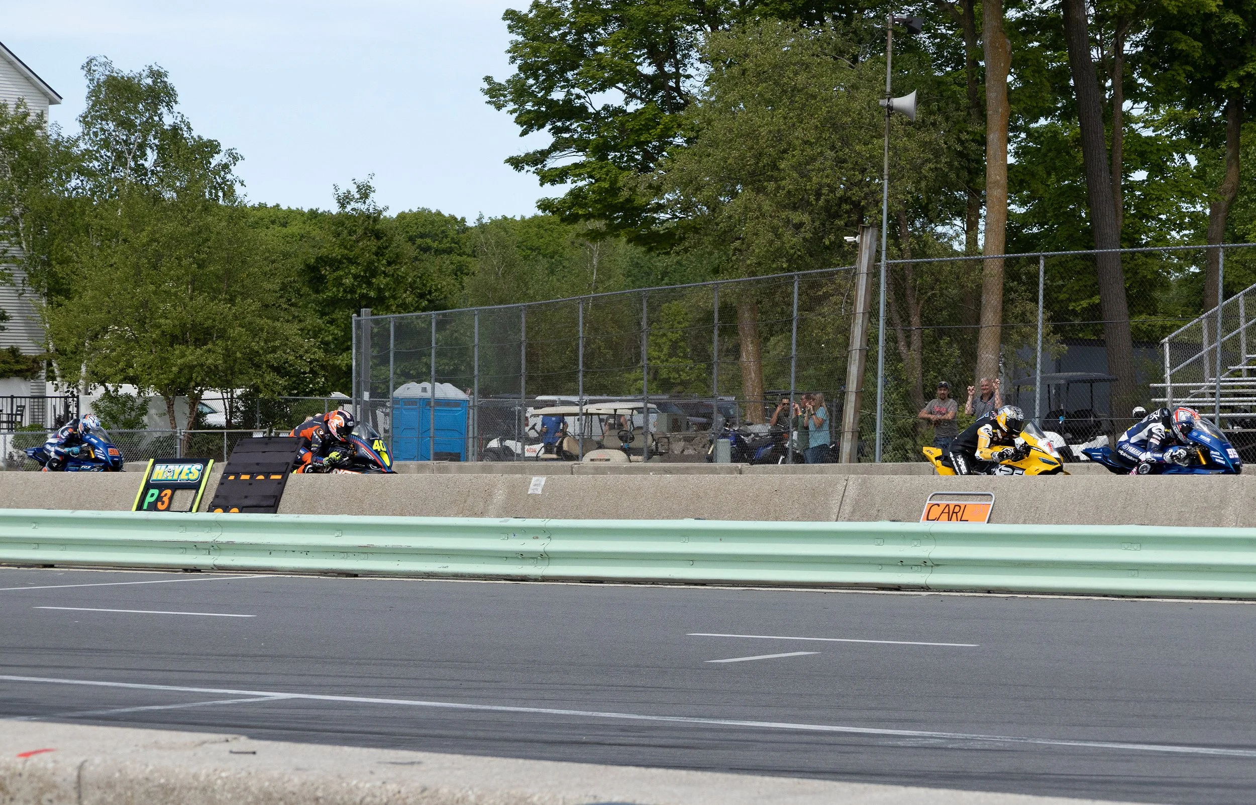 Motorcycles racing on a track with spectators behind a fence and tall trees in the background.