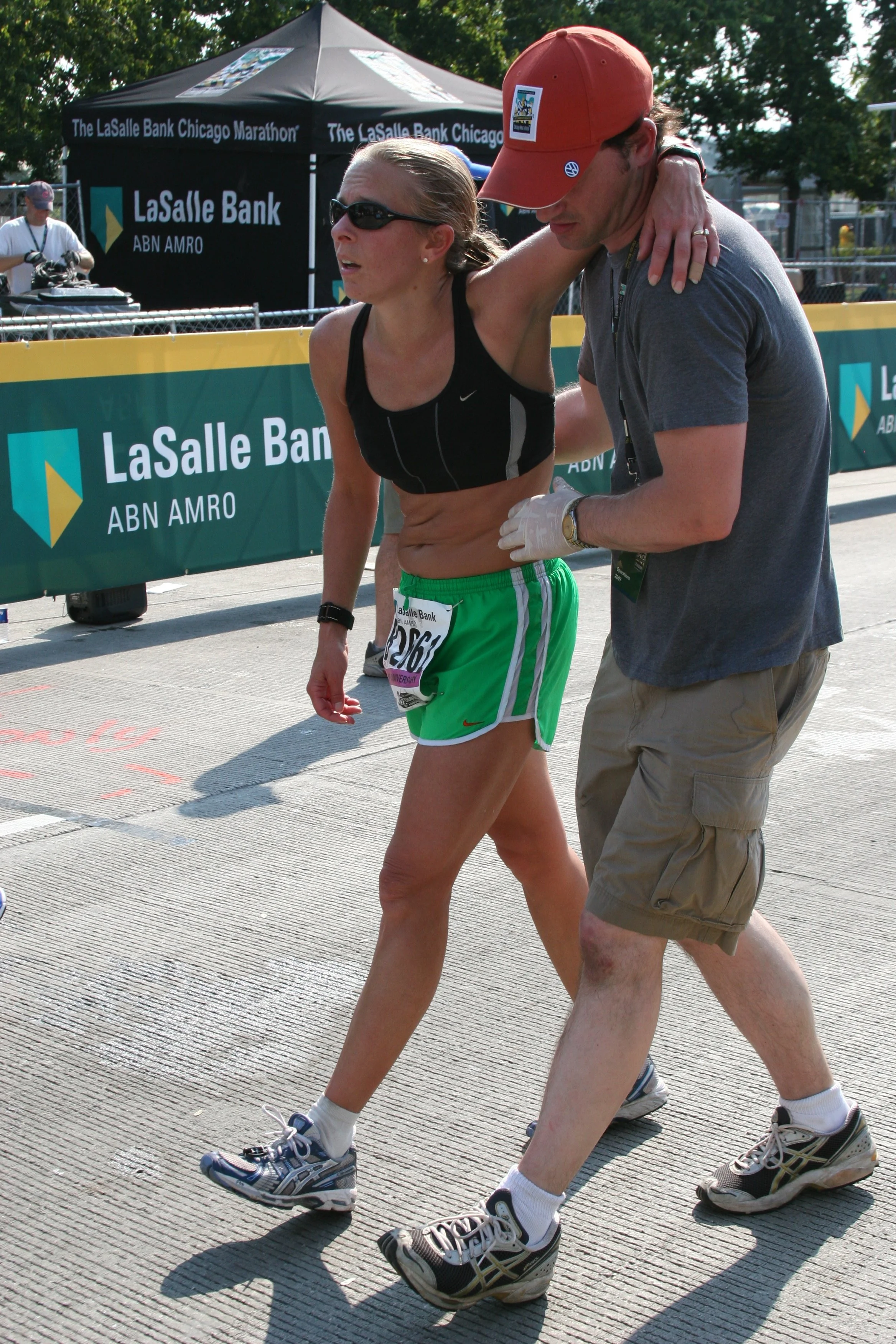 A woman with blonde hair, wearing sunglasses, a black tank top, and green running shorts, being assisted by a man wearing a red baseball cap, grey T-shirt, and khaki shorts, during a marathon.