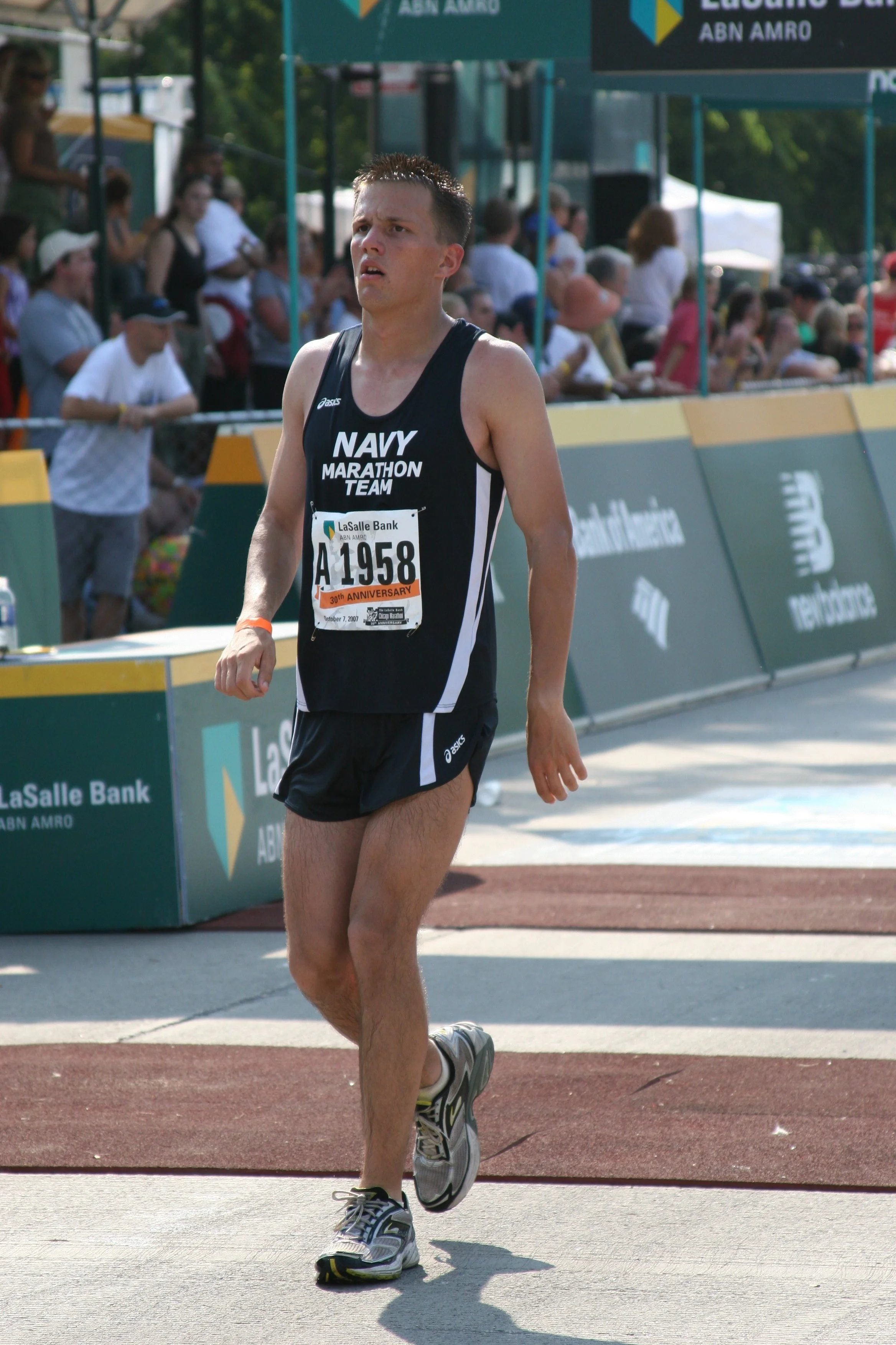 A male marathon runner wearing a black tank top with white lettering that reads 'NAVY MARATHON TEAM' on the front and a race bib number A 1958, walking on a track near the finish line of a marathon event. The background shows spectators behind a fenc