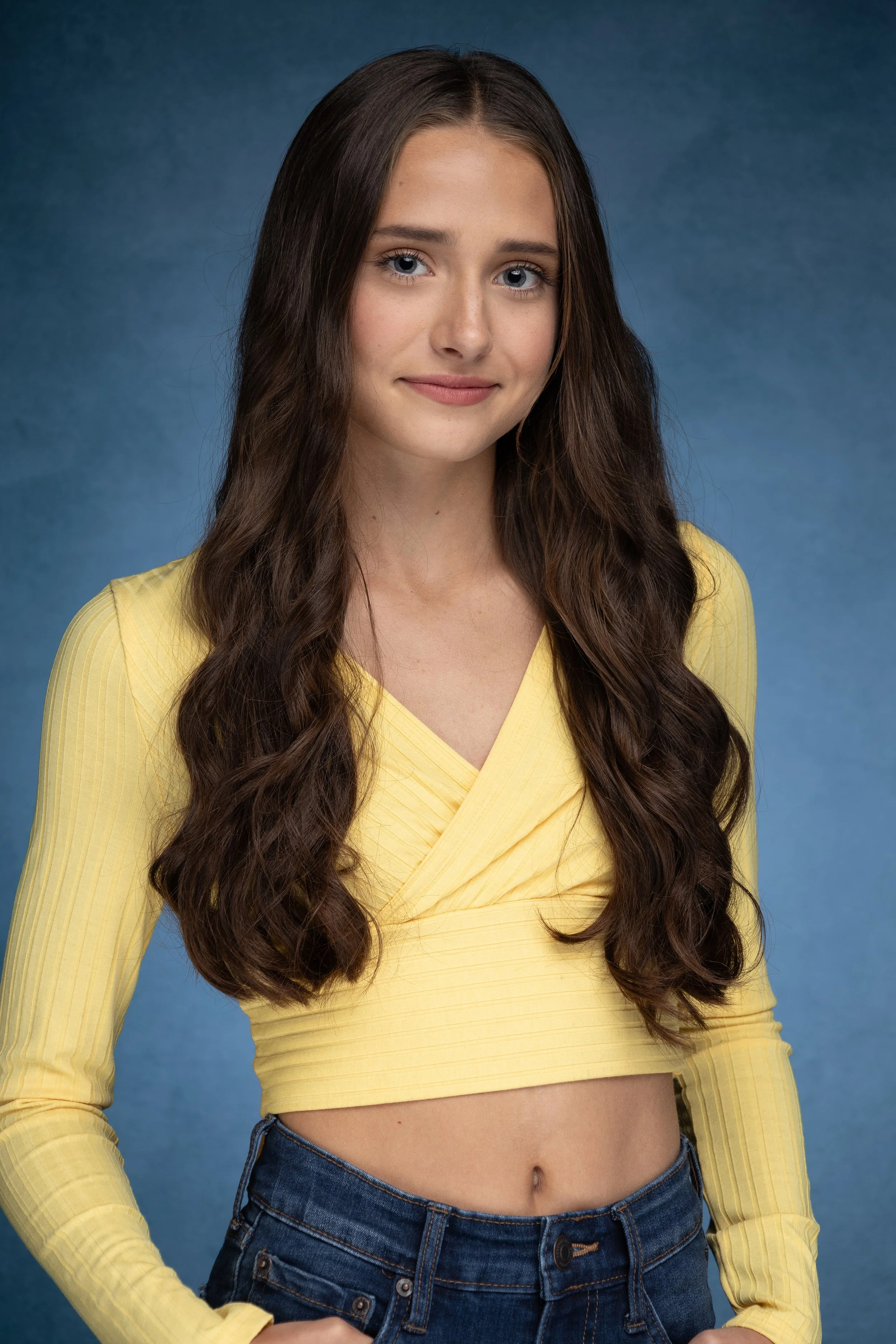 A young woman with long wavy brown hair wearing a yellow long sleeve crop top and jeans, posing against a blue background.