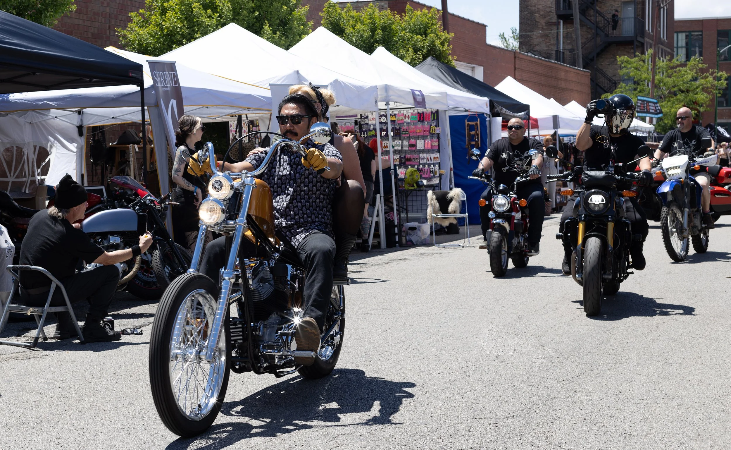 A street scene at an outdoor market with white market tents. Several motorcyclists riding bikes are passing by, with one rider in the foreground wearing sunglasses and a patterned shirt. Market stalls behind them display various items, and people are