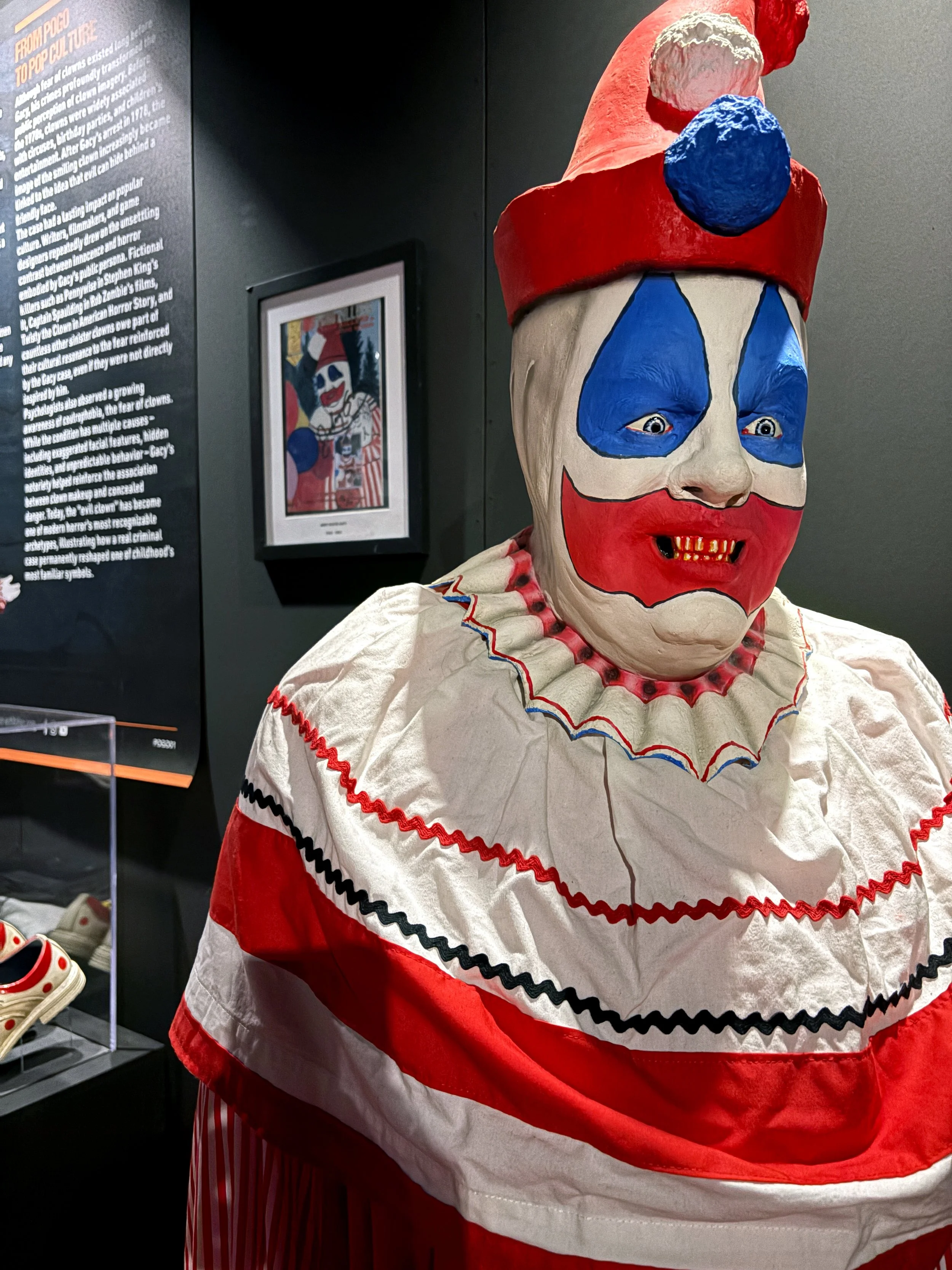 A clown costume with a large, painted clown mask featuring exaggerated red lips, blue makeup around the eyes, a red nose, and a red and white hat with a blue pom-pom on top. The costume includes a red and white ruffled outfit with decorative stitchin