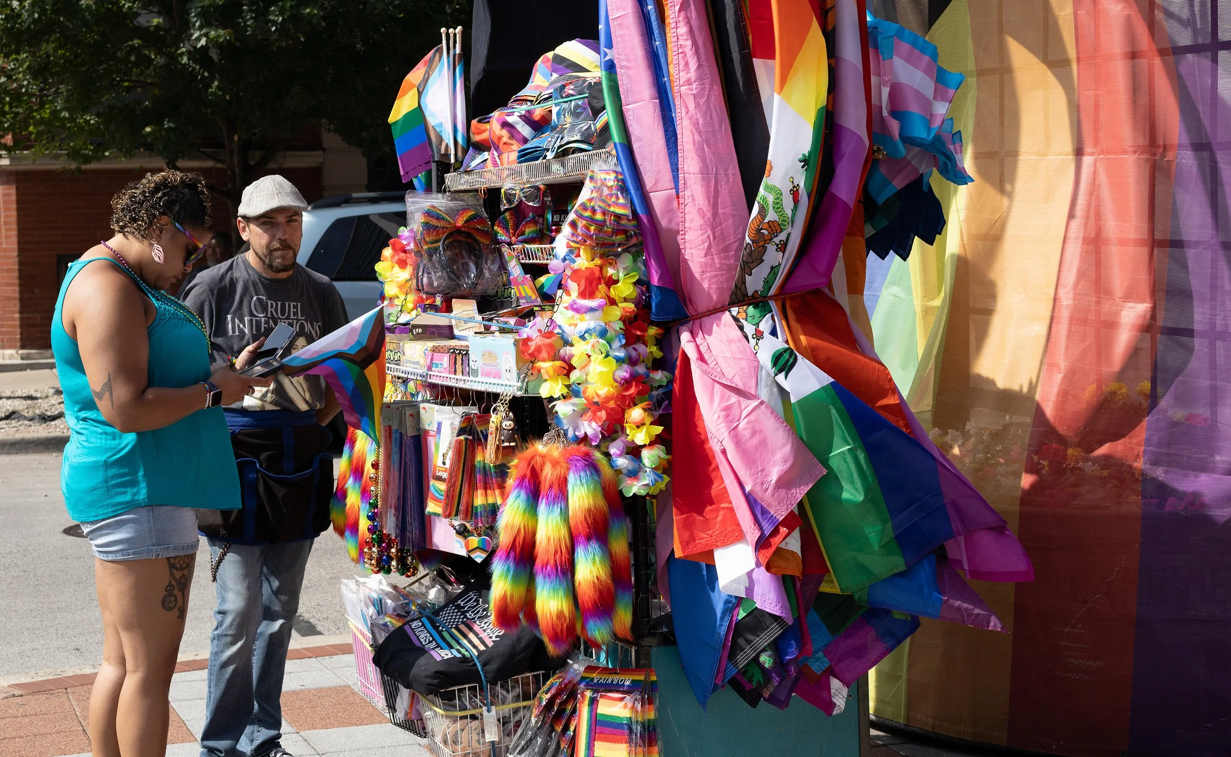 A person with curly hair and colorful earrings is looking at their phone while standing in front of a street vendor's booth that sells rainbow-colored flags, clothing, and accessories decorated for a Pride celebration. A man beside them is wearing a 