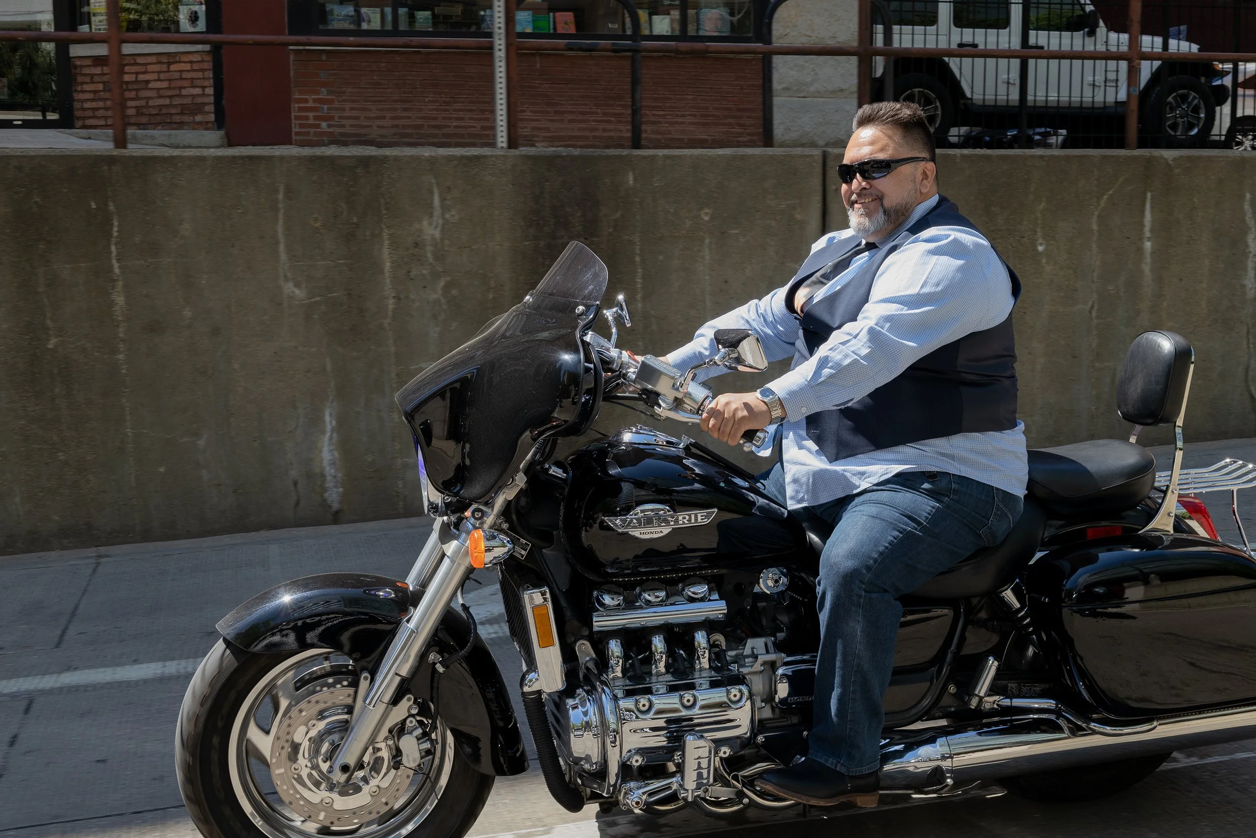 Man with sunglasses wearing a dress shirt and vest riding a black motorcycle on city street.