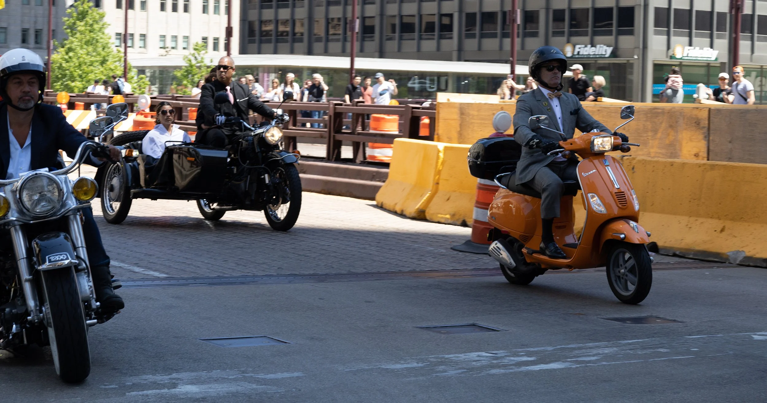 Three men riding motorcycles and a scooter on a city street, with pedestrians and office buildings in the background.