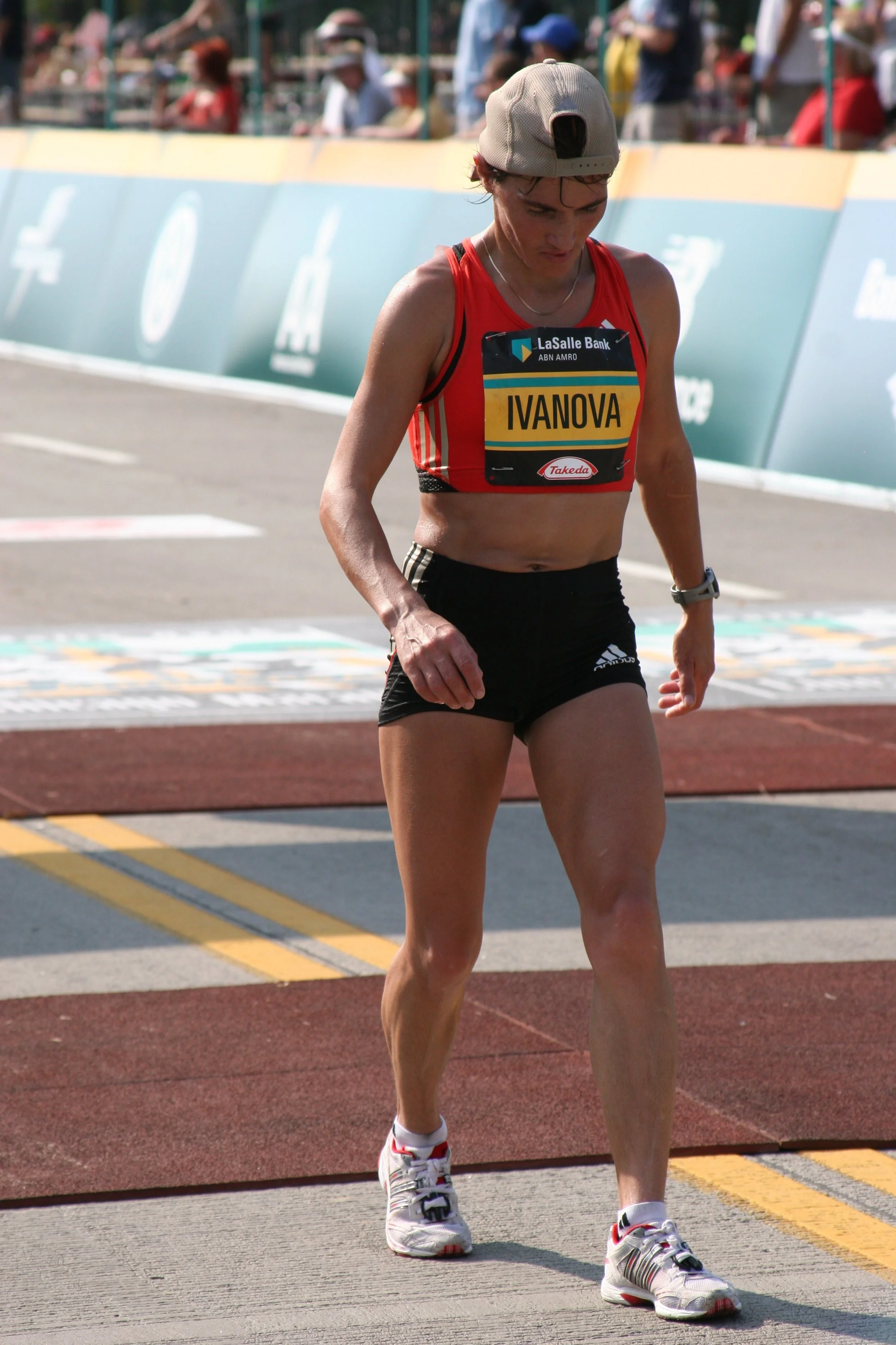 A female athlete in a red running tank top and black shorts walking on a track during a race, with spectators in the background.