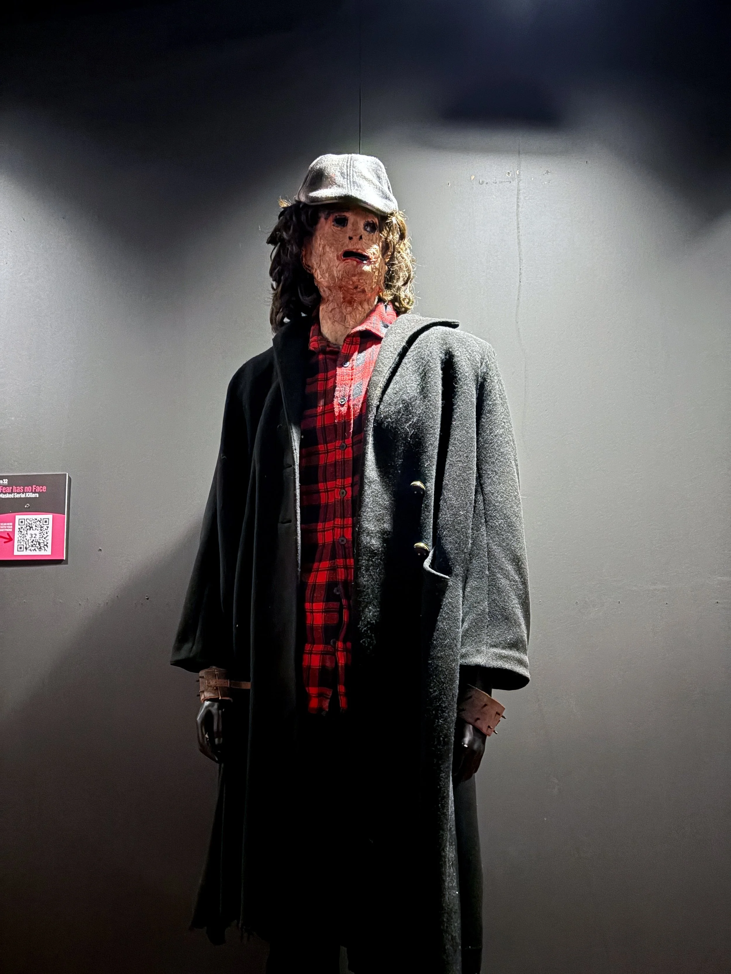 A mannequin dressed in a gray coat, red plaid shirt, black gloves, and a gray hat, with a gray coat draped over its shoulder, standing against a plain gray wall in a museum exhibit.