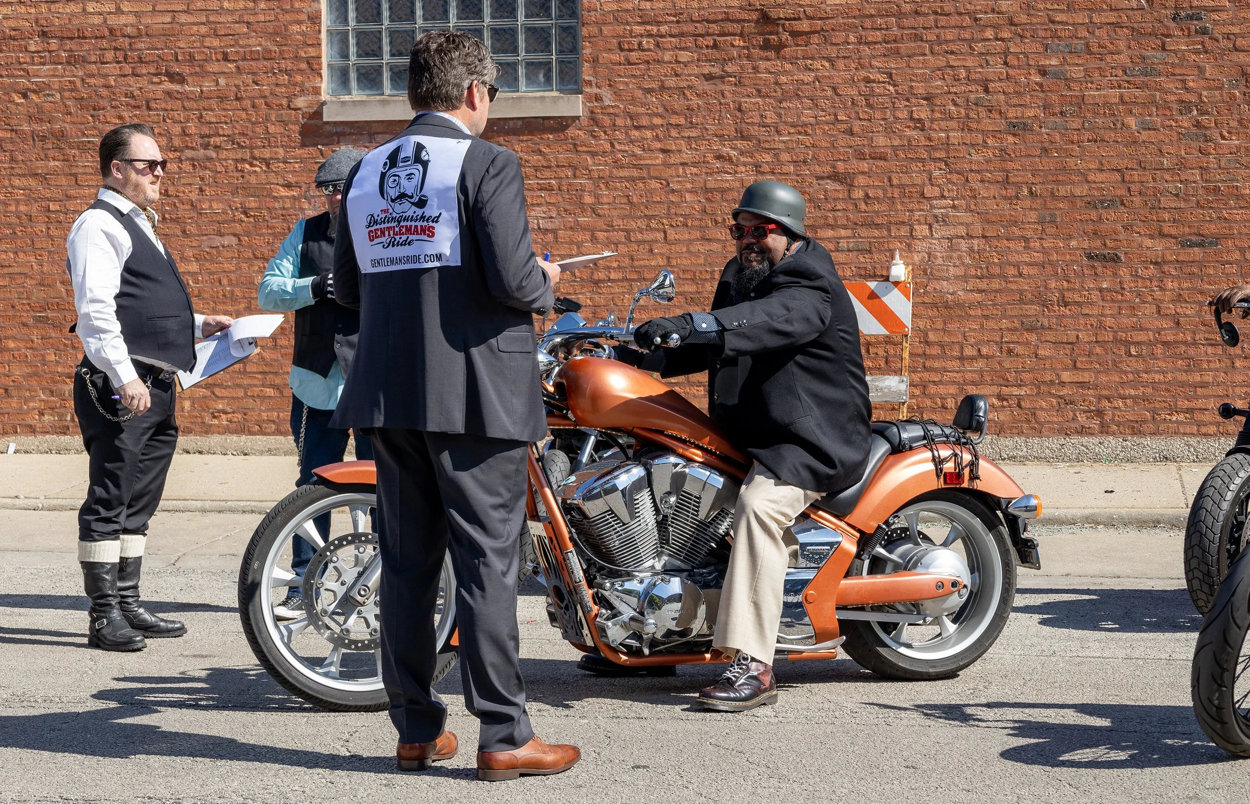 A man sitting on an orange motorcycle, wearing a black jacket, beige pants, red sunglasses, and a black helmet, talking to a man in a suit with a notepad. Several other men stand nearby, one holding papers, outside in front of a brick building.