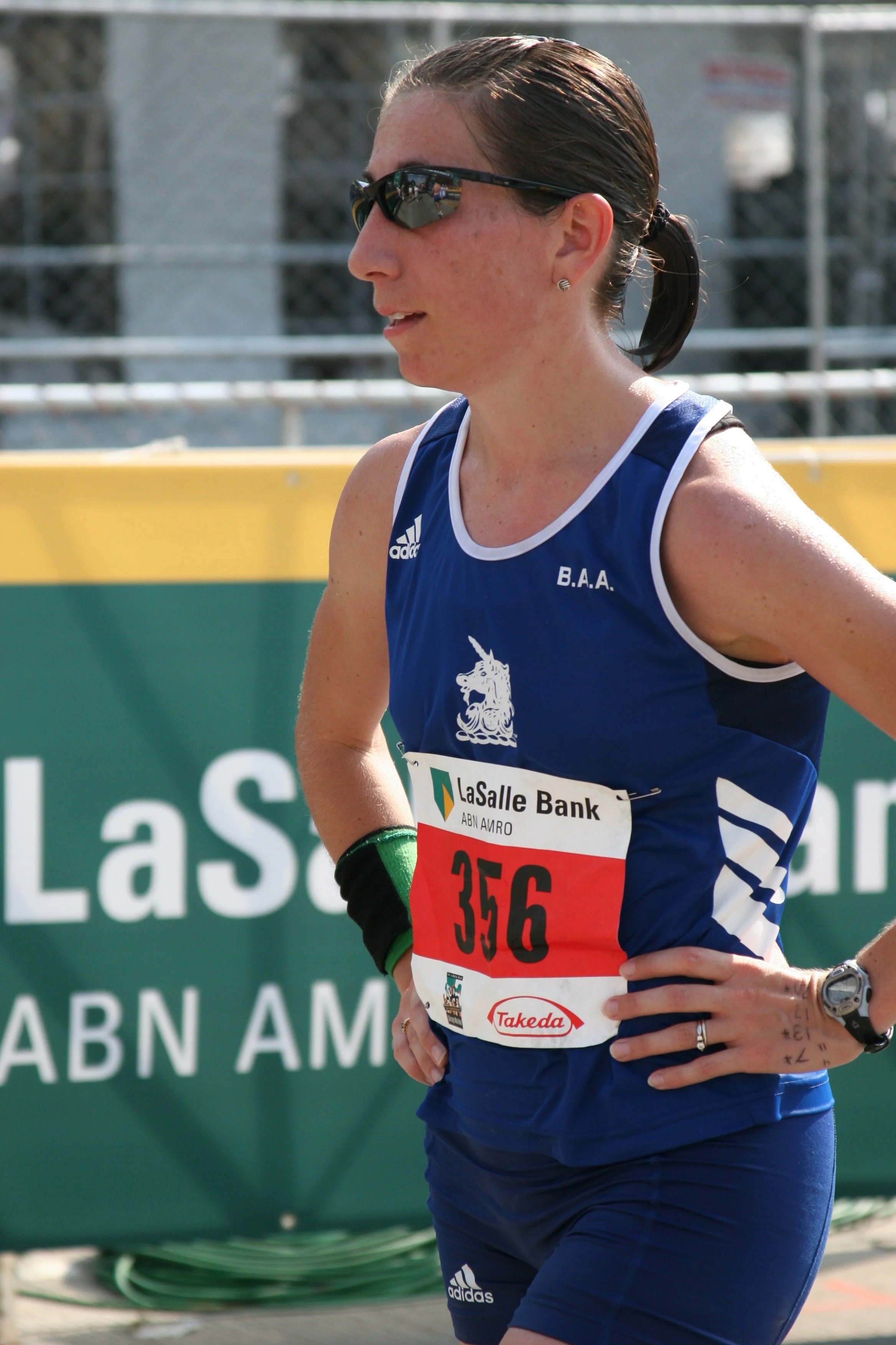 Close-up of a female runner wearing sunglasses, a blue sports jersey with white accents and a red race bib, standing outdoors near a green fence.