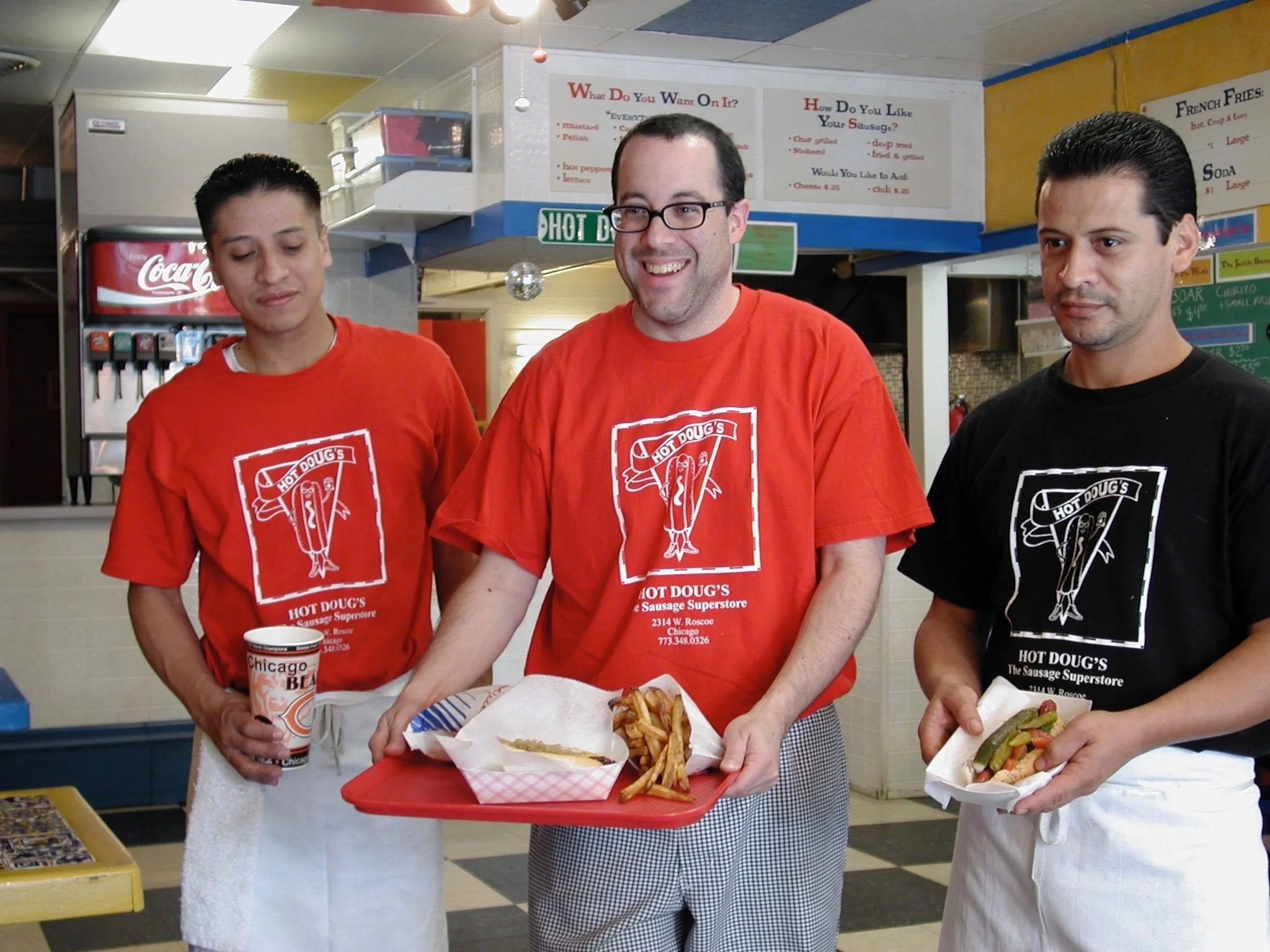 Three men in red and black T-shirts serving food at a counter in a fast-food restaurant, with menus and soda dispensers in the background.