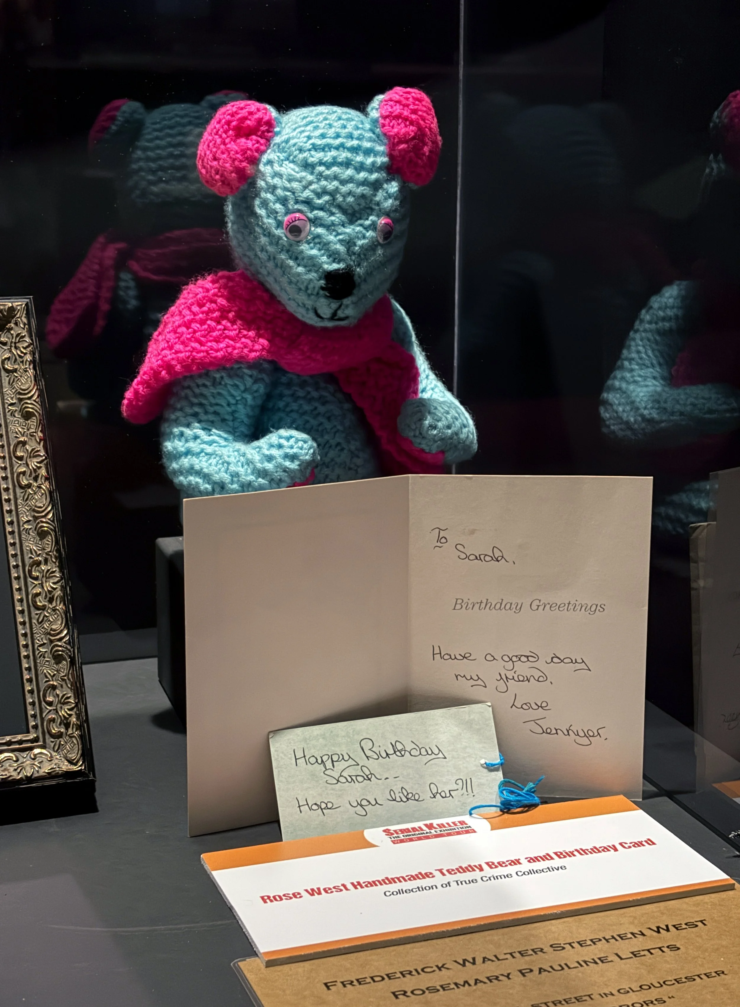 A handmade crocheted teddy bear with pink ears and a pink scarf, displayed behind a family birthday card on a black surface.