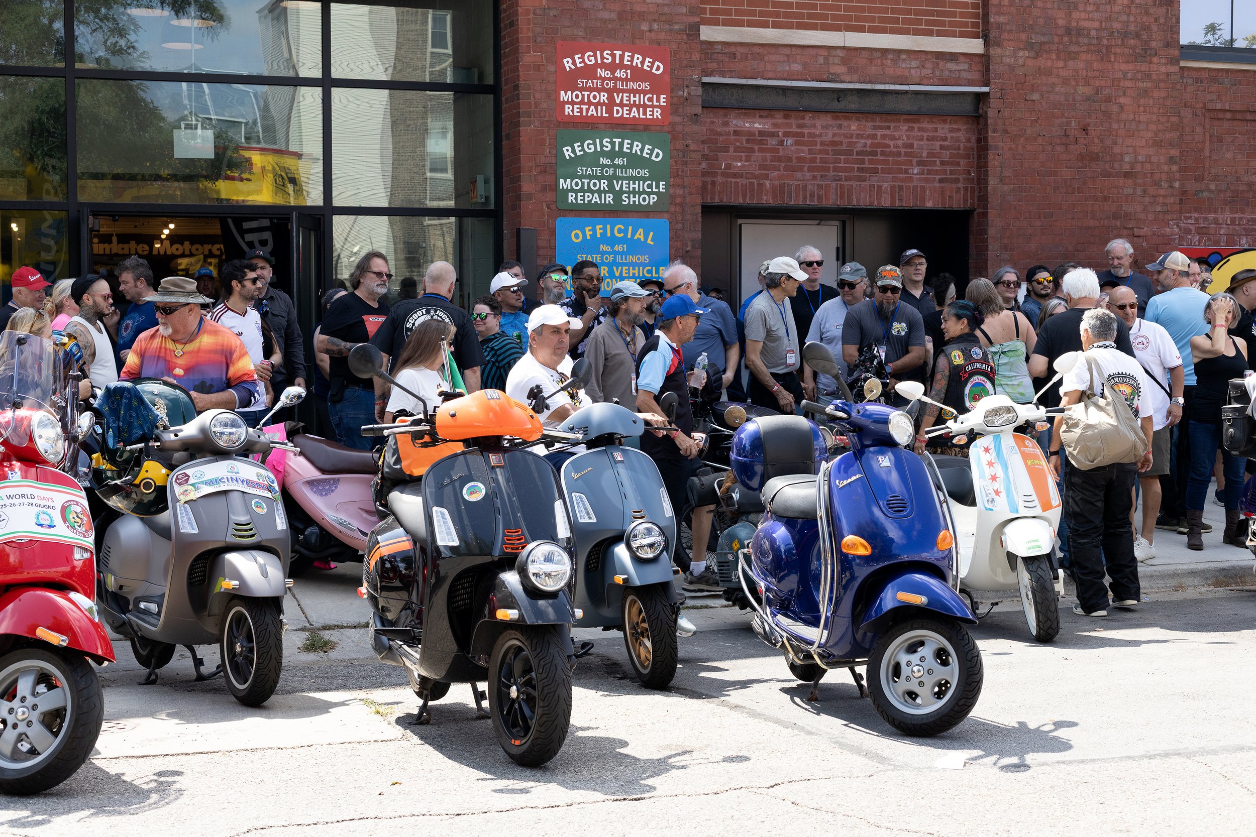 A large group of people gathered outside a building, with many scooters parked in the foreground. The building has signs indicating it is a recognized motor vehicle dealer and repair shop in Illinois.