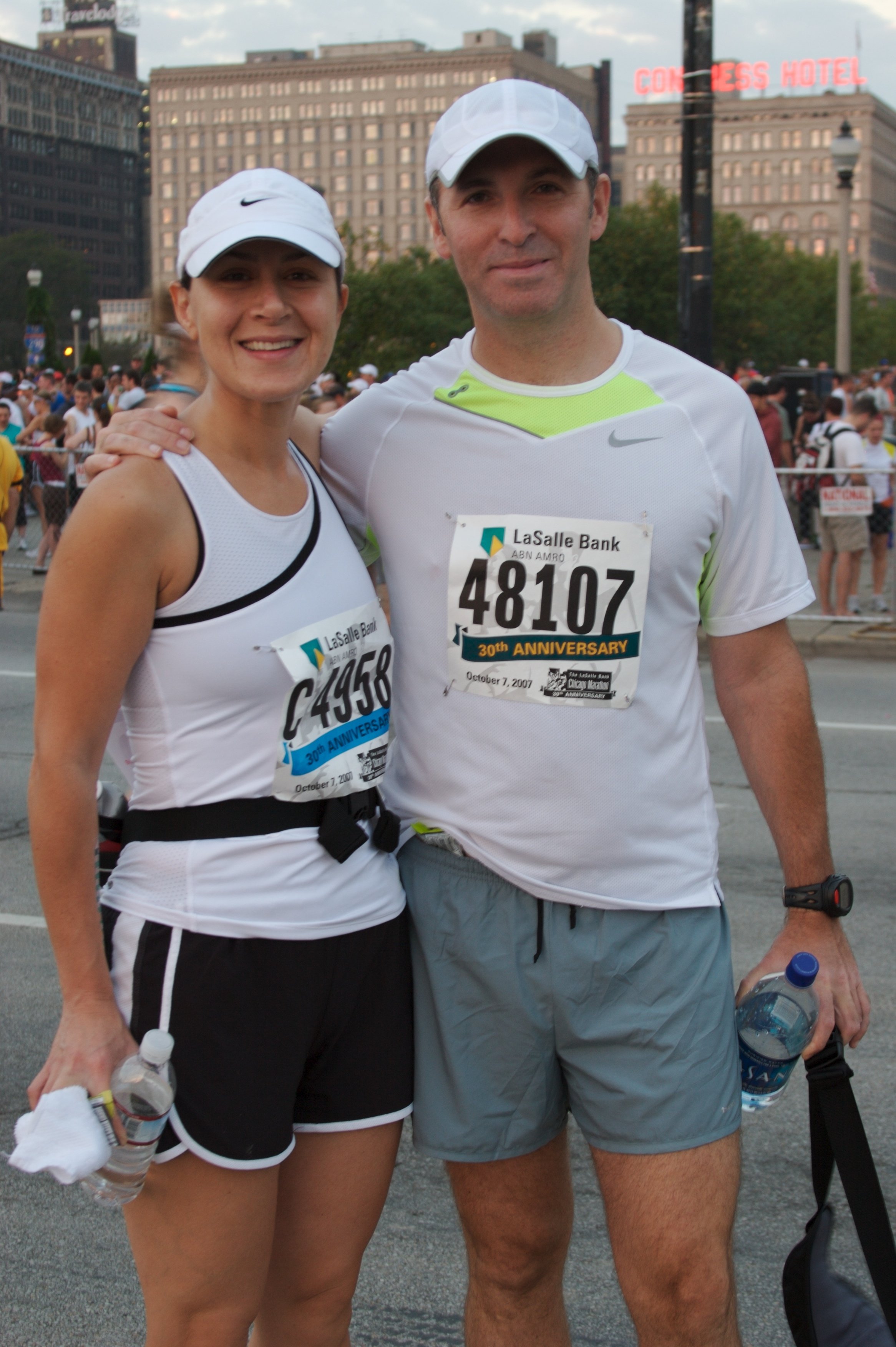 Two marathon runners posing for a photo, smiling, with a crowd and city buildings in the background. The woman is wearing a white cap, a white tank top, and black shorts, holding a water bottle. The man is wearing a white cap, a white athletic shirt 
