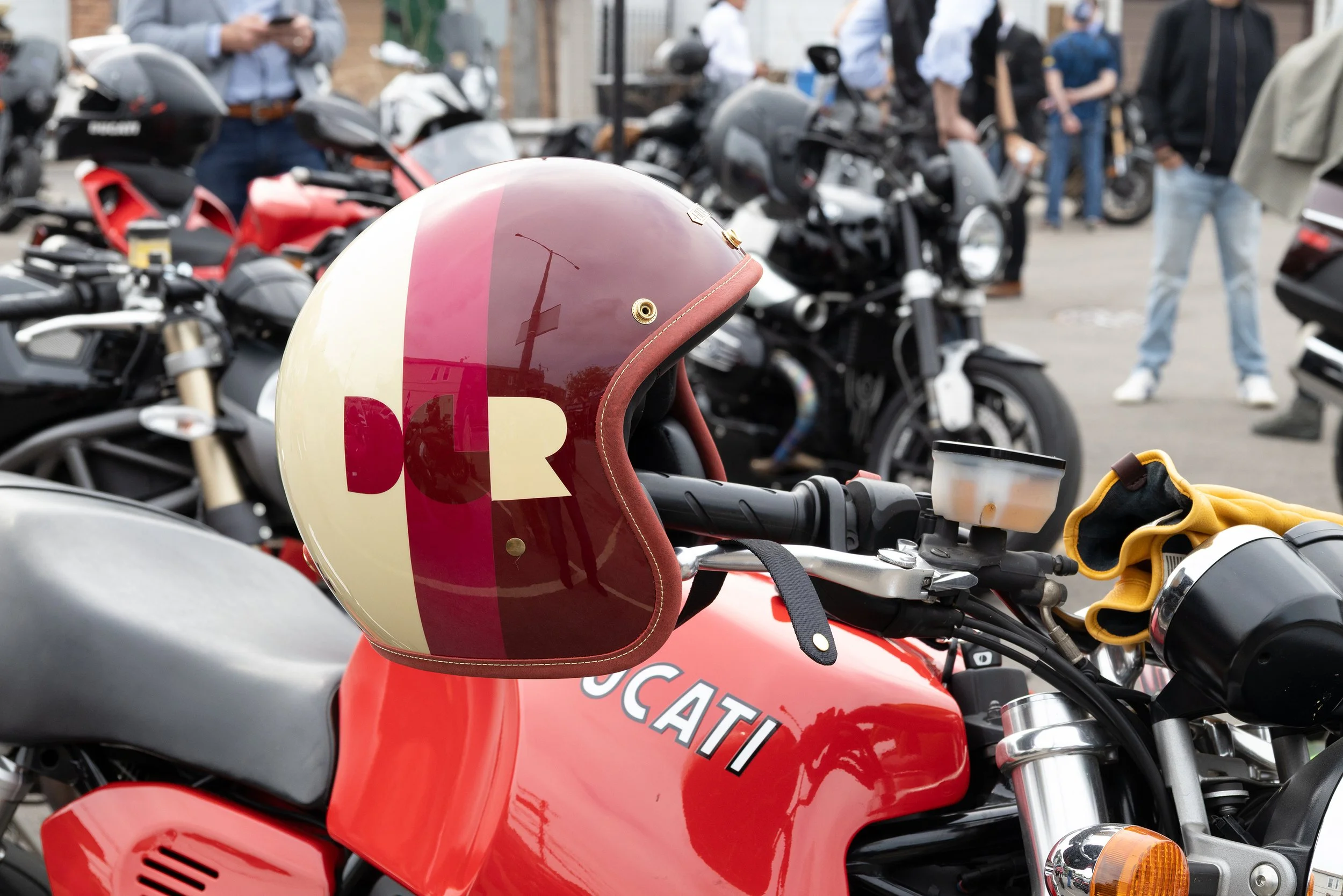 Close-up of a red motorcycle with a white and pink helmet resting on the handlebar. Background shows multiple motorcycles and people standing around.