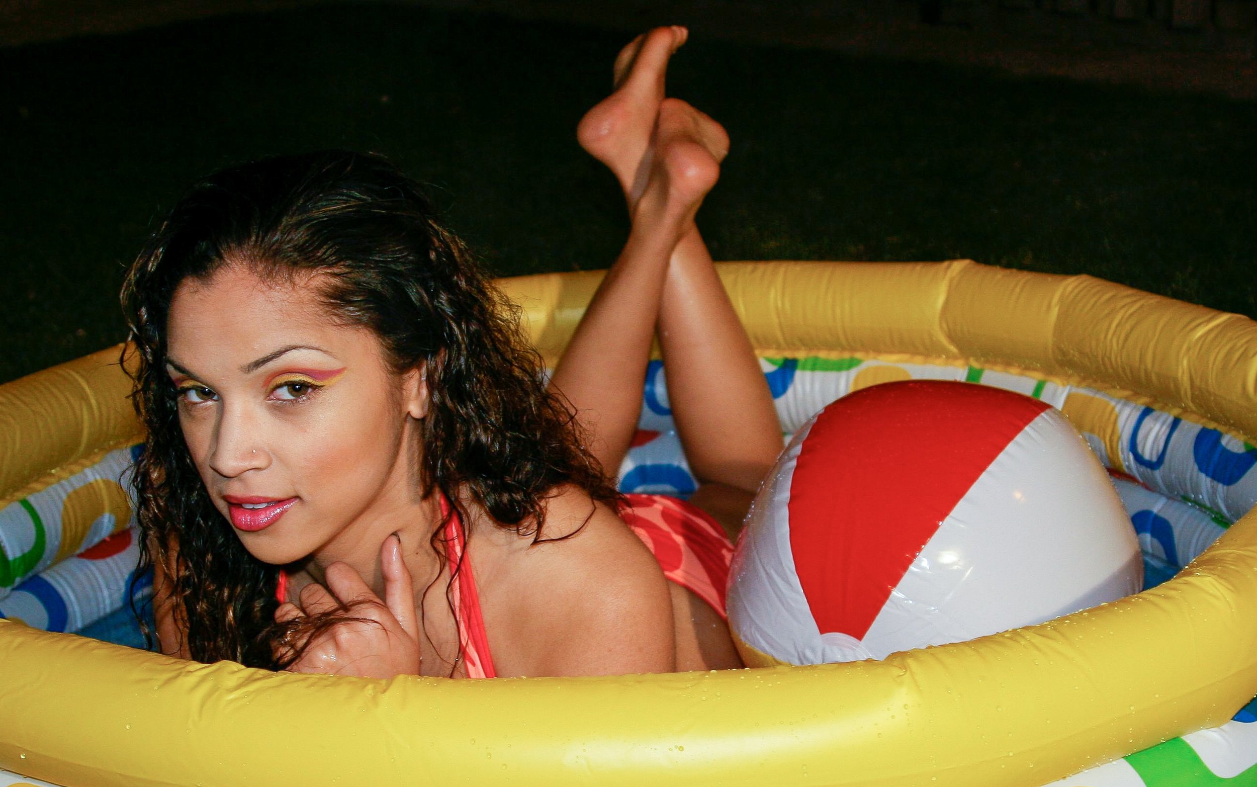 A woman with curly dark hair and colorful eye makeup lying in a yellow and blue inflatable pool with a red and white beach ball. It is nighttime or in a dark setting.