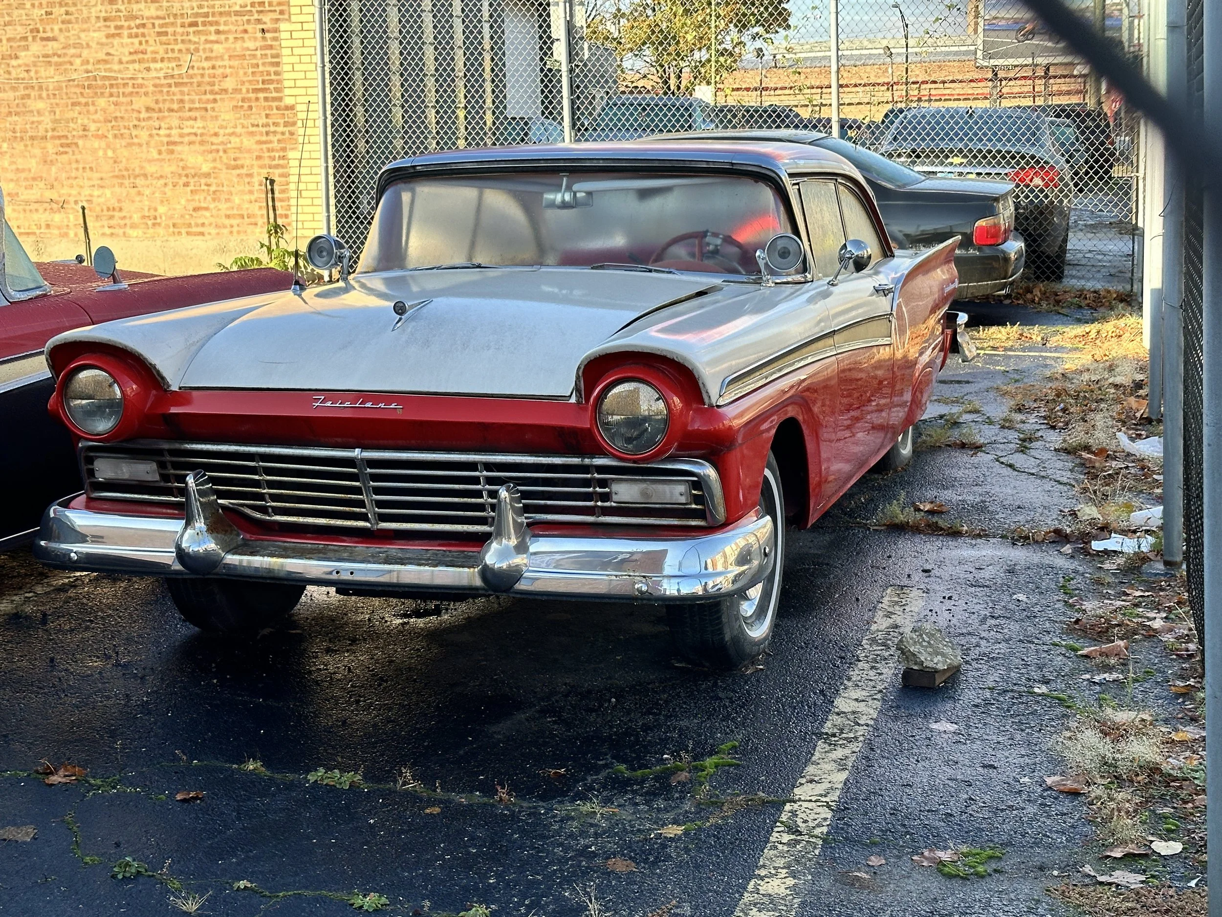 A vintage car, primarily silver with red accents, parked in an alleyway with a chain-link fence in the background. The car has prominent tail fins and round headlights, with some speaker devices attached to the front. There is another black car parke