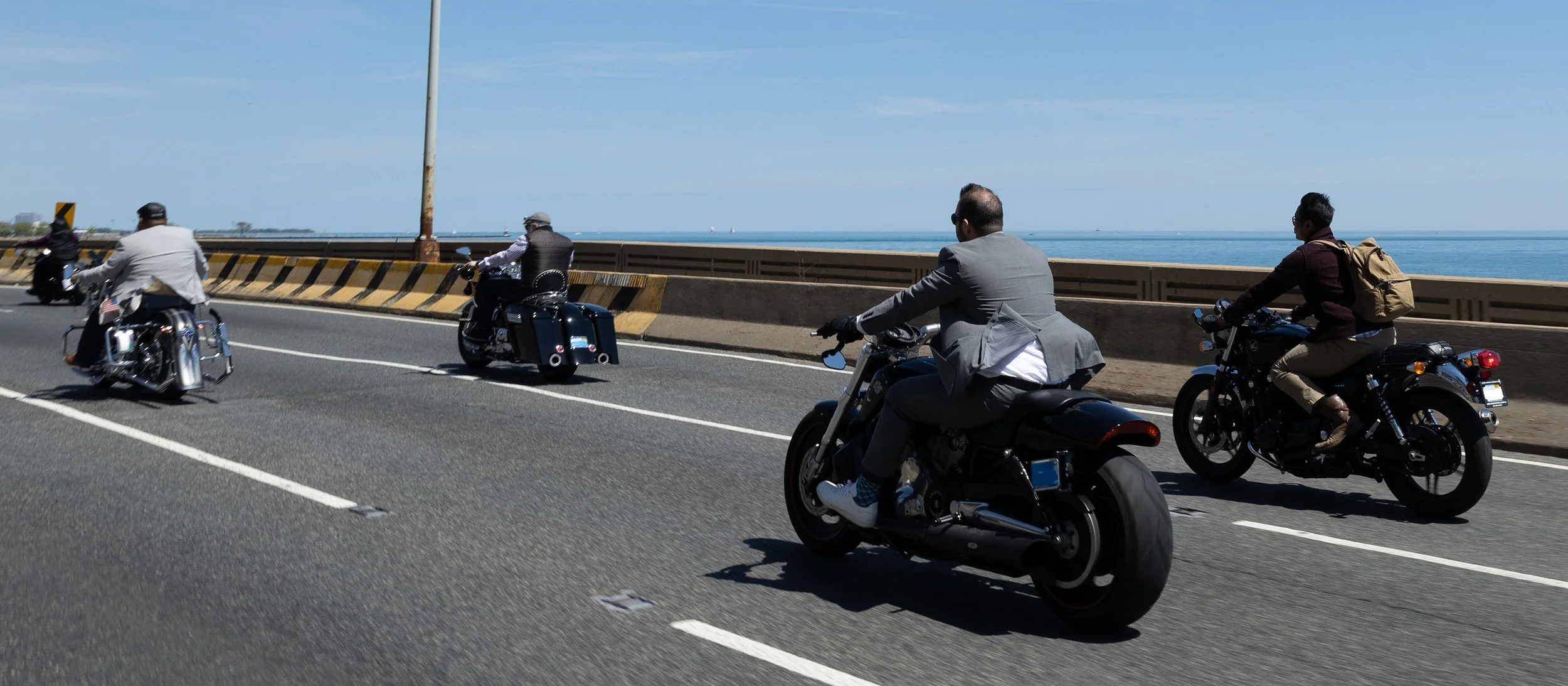 Four motorcyclists riding on a highway beside a body of water on a clear day, some dressed in business attire and casual clothing.