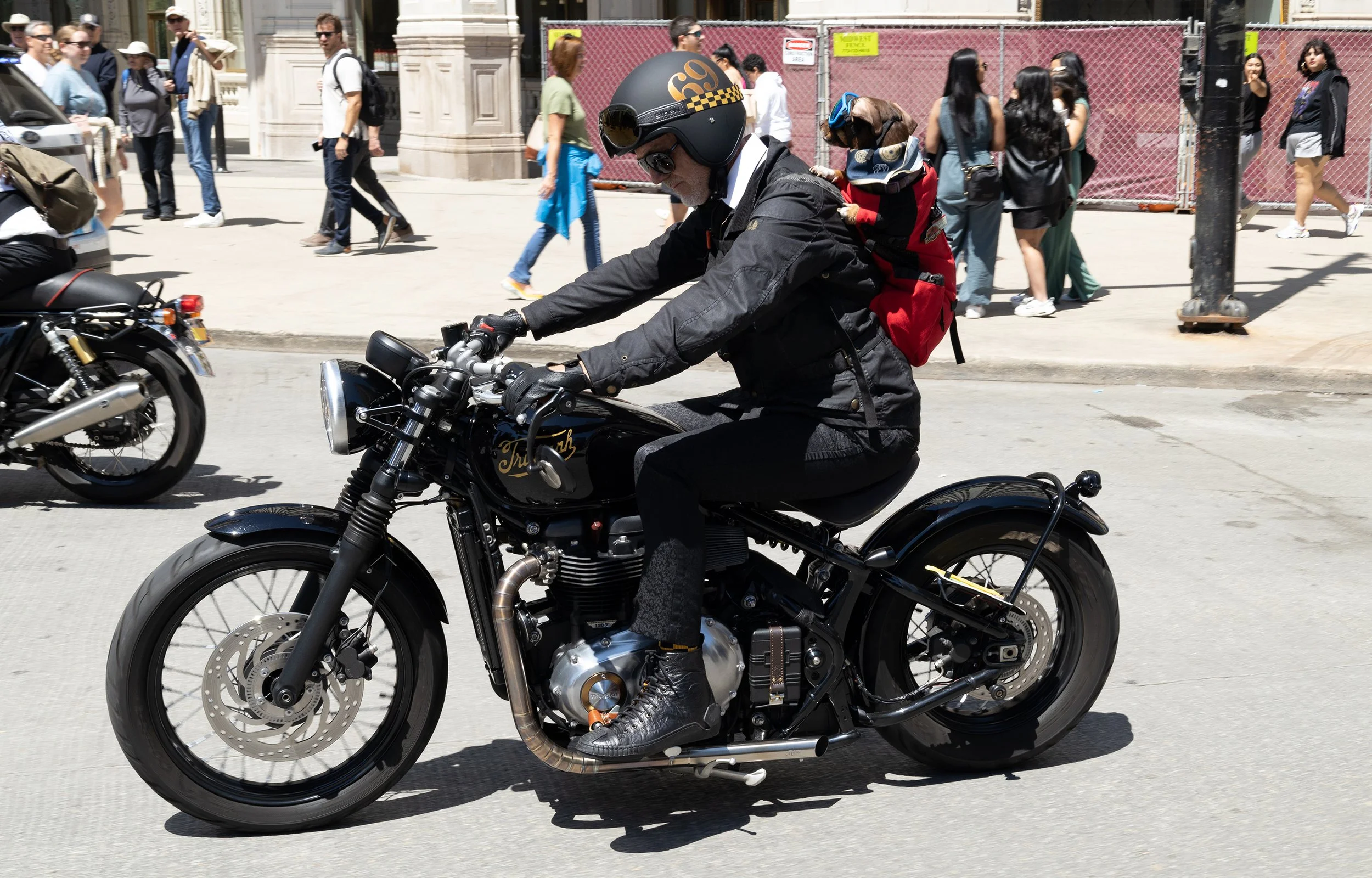 A man wearing a black jacket, black helmet with a yellow checkered stripe, sunglasses, and black gloves riding a black Triumph motorcycle on a city street. Several pedestrians walk in the background, some with backpacks, near a construction fence and