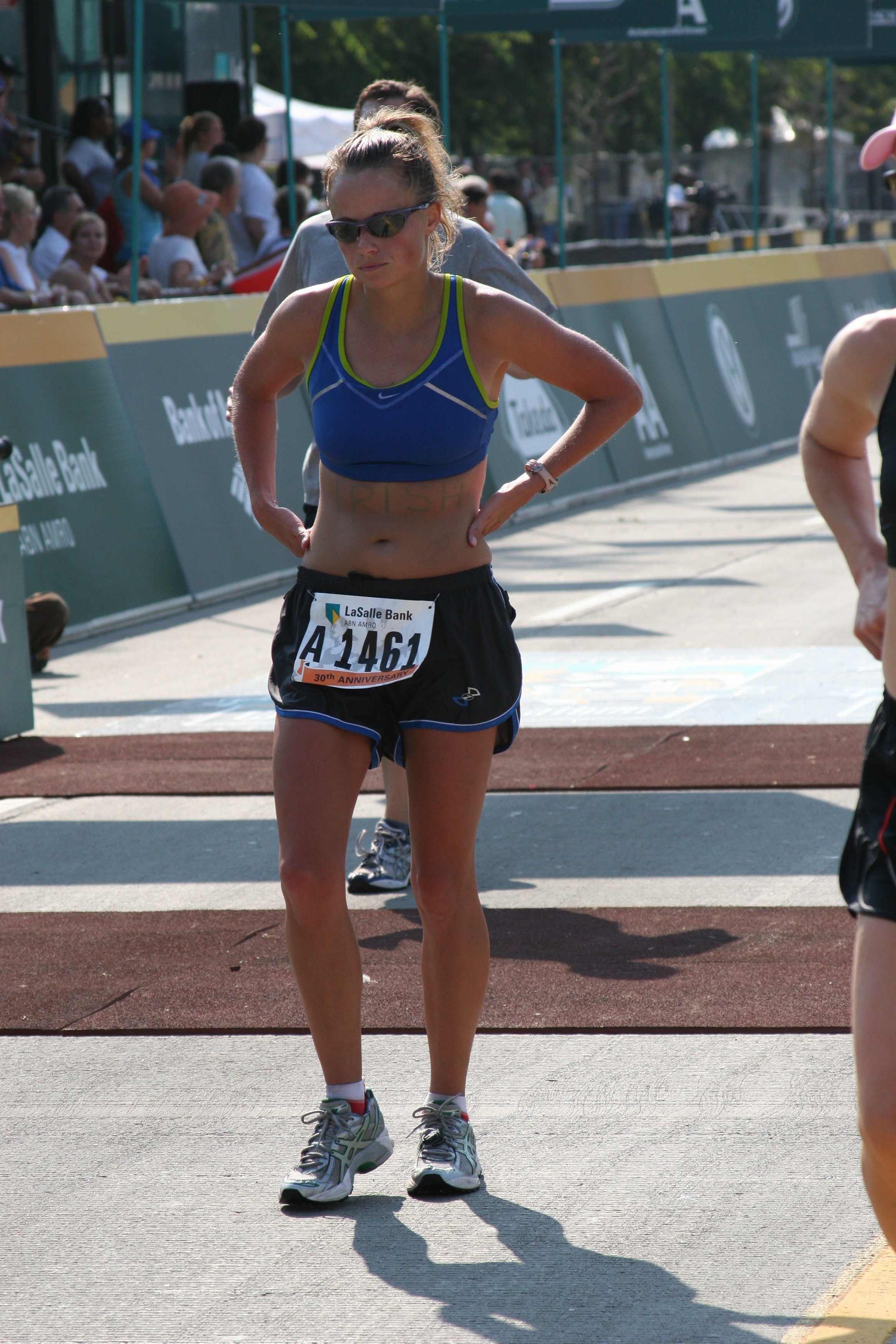 Female athlete wearing sunglasses and a blue sports bra, standing with hands on hips at a race event on a sunny day.