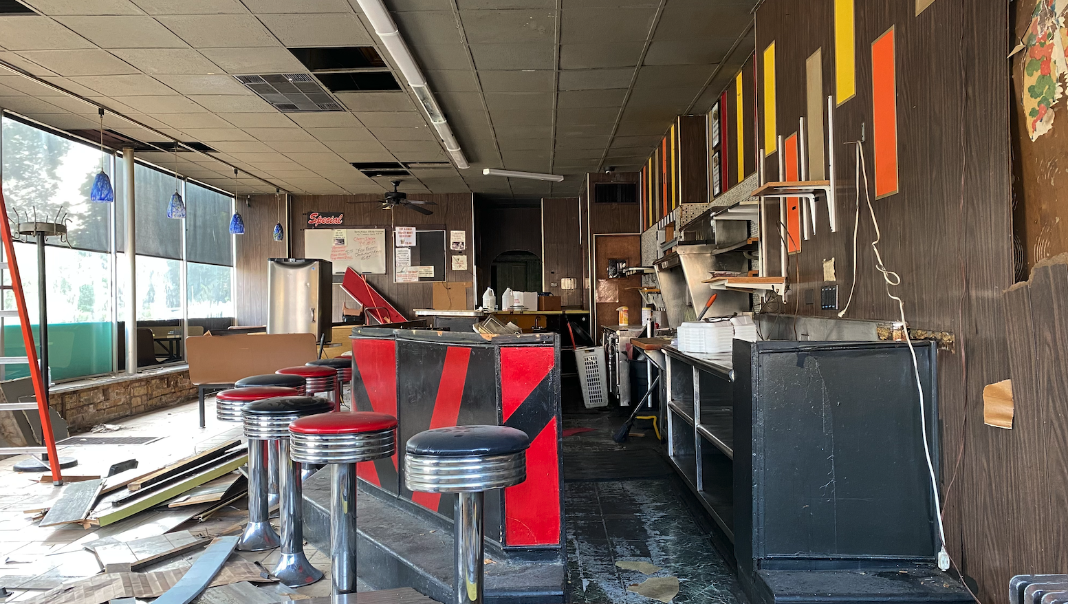 Interior of a closed pizza restaurant with peeling floors, damaged ceiling, and disorganized furniture and supplies, indicating it is abandoned or under renovation.