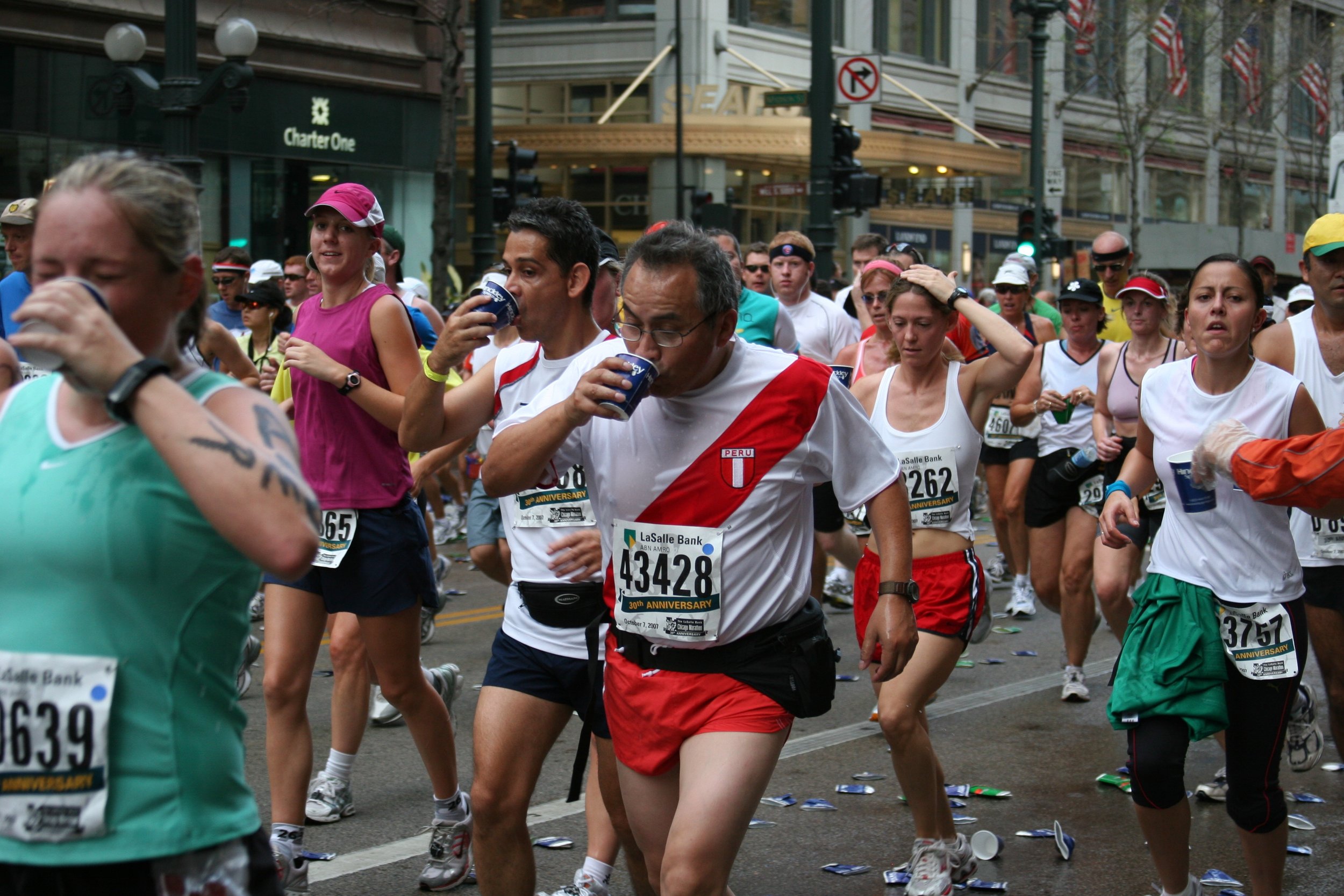 Crowd of marathon runners participating in a street race, with some drinking water and others focused on running. Debris is scattered on the street, and buildings with American flags are in the background.