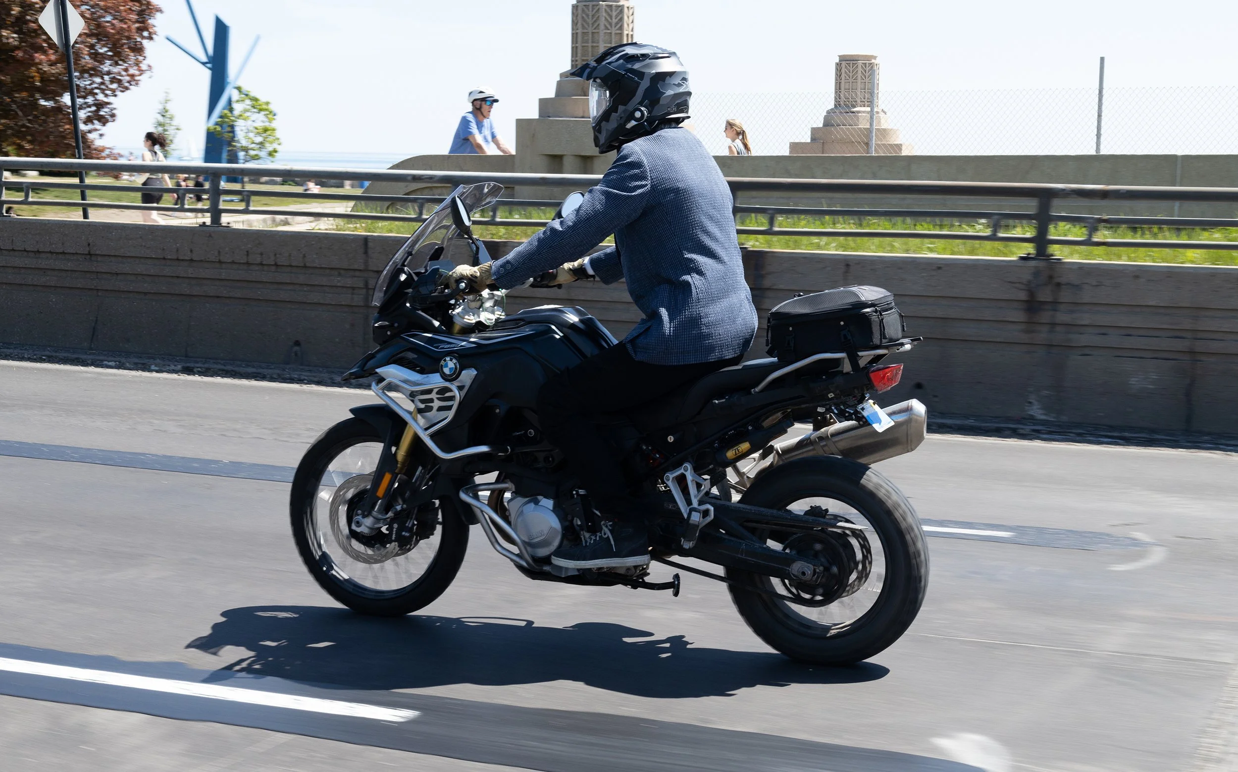 A person wearing a helmet and a blue jacket riding a black motorcycle on a city street. In the background, there are pedestrians, a metal fence, a chain-link fence, and some tall structures.