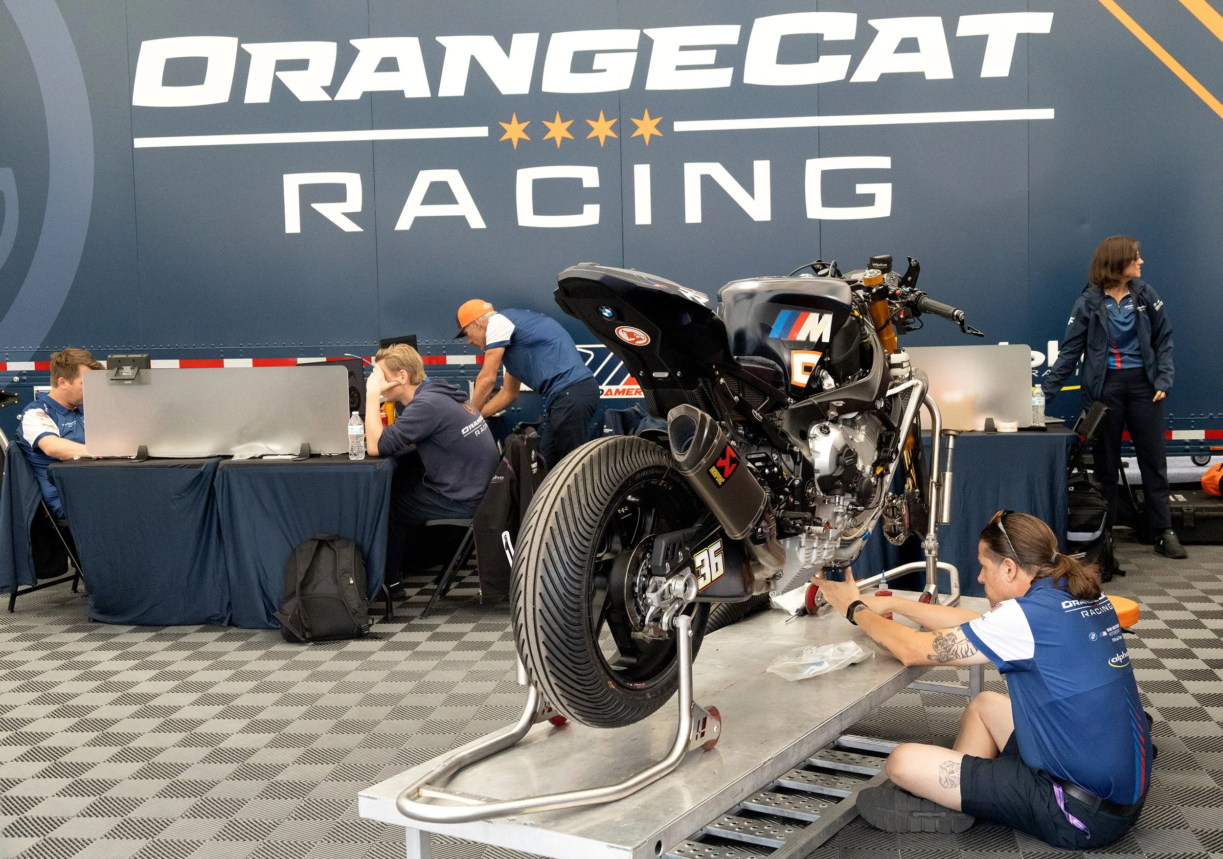 A motorcycle and several team members working in a garage at Orange County Racing, with a large blue wall featuring the event's logo and name.