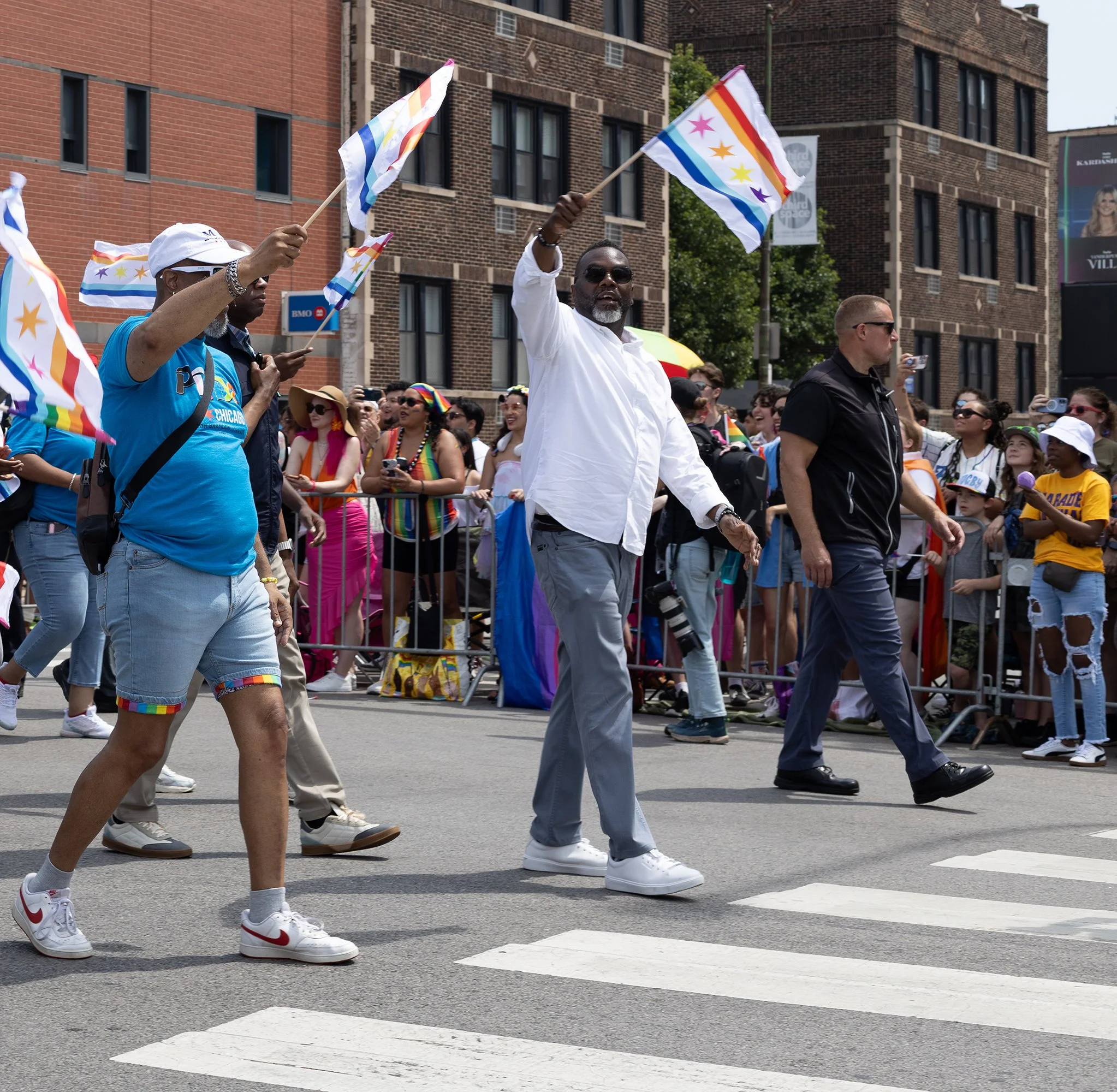 People participating in a pride parade, holding colorful pride flags and walking on the street with crowd and buildings in the background.