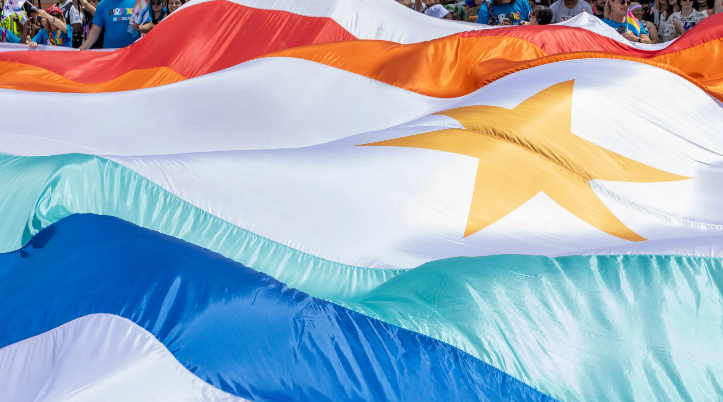 Crowd of people at an outdoor event holding a large rainbow flag with a yellow star in the center.