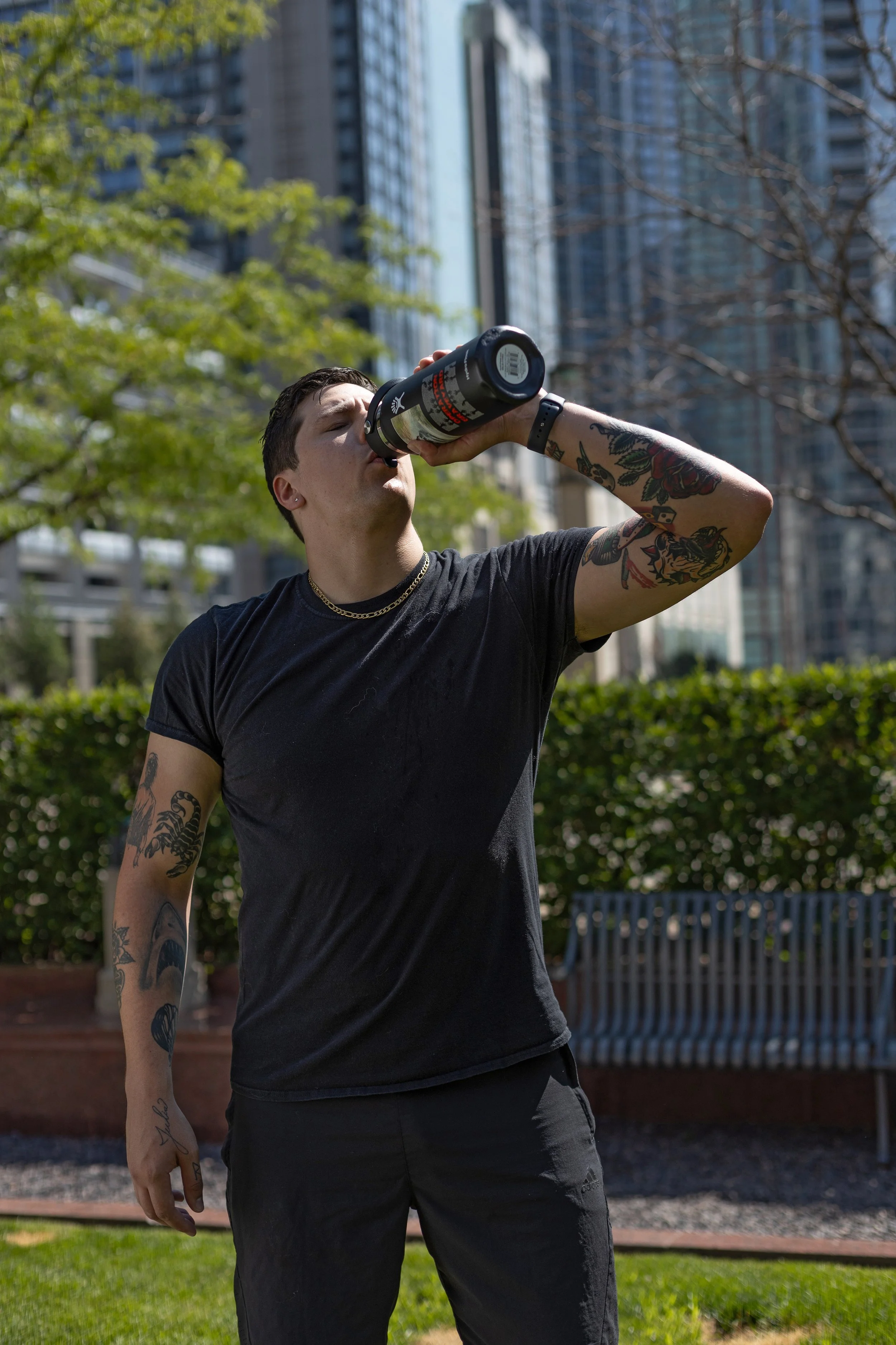 A man with tattoos on his arm drinking from a black insulated water bottle outdoors with tall buildings and trees in the background.