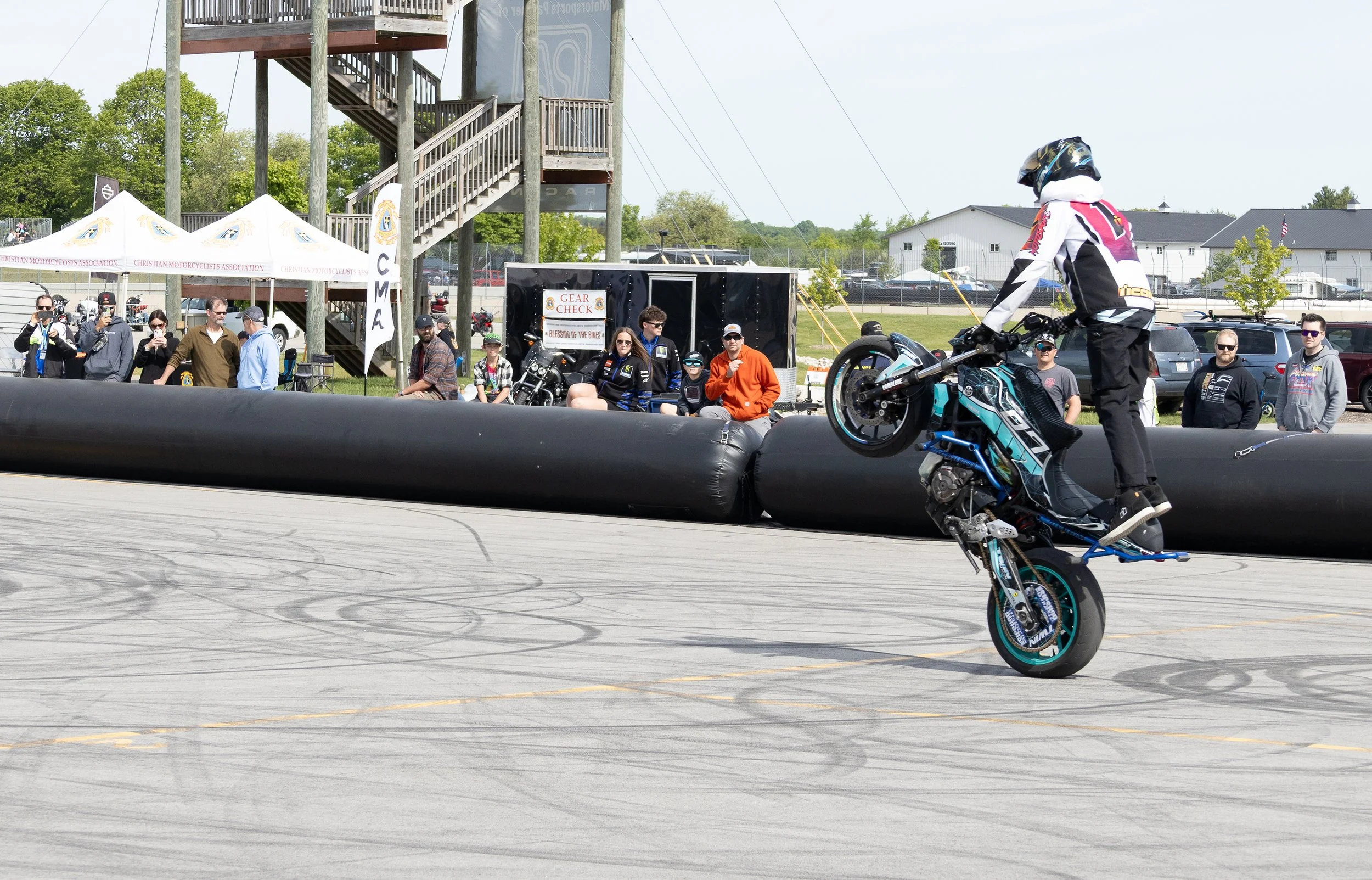 A motorcycle stunt rider performing a wheelie on a blue and black motorcycle at a motorcycle event, with spectators watching and taking photos in the background.