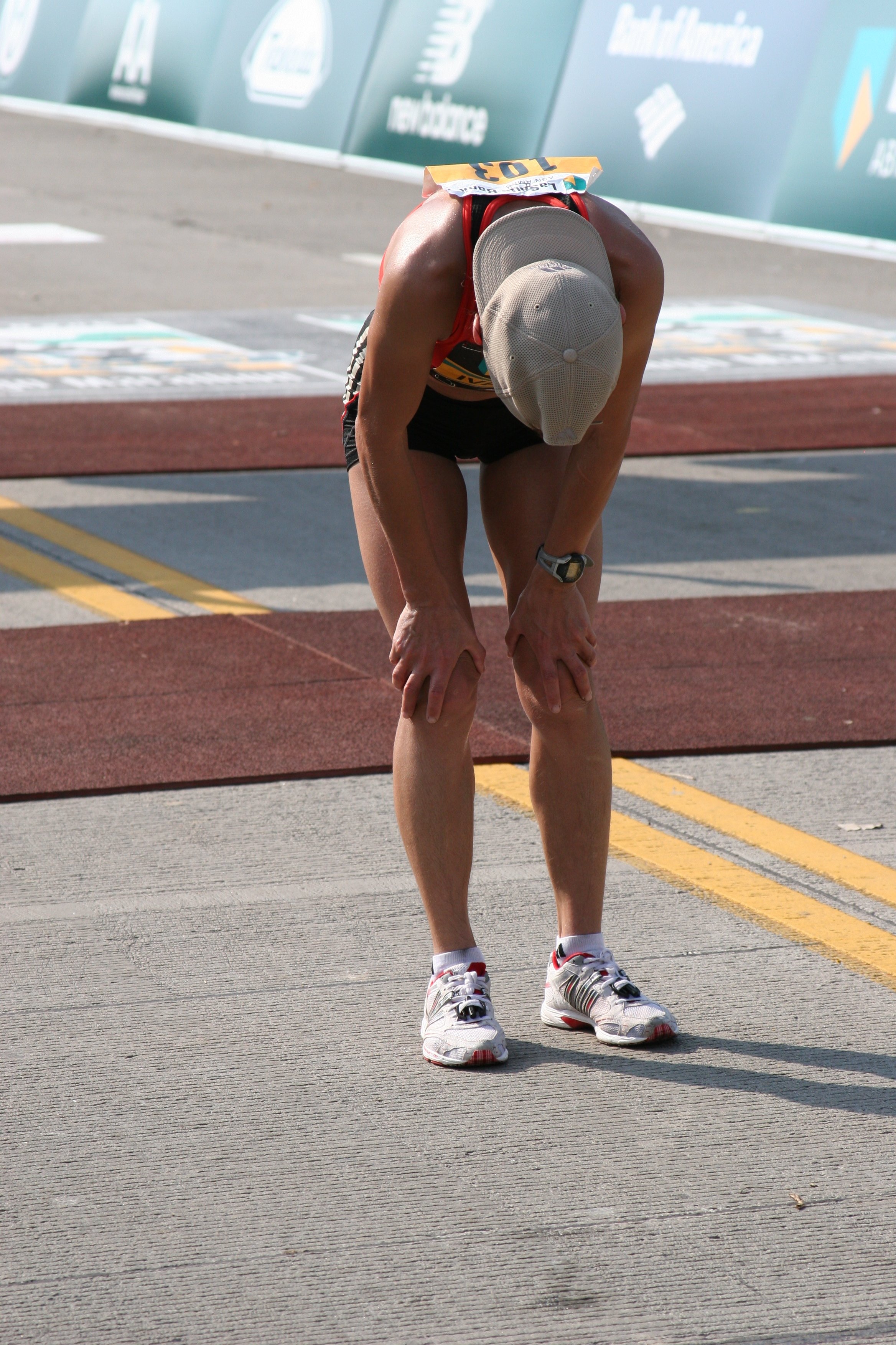 A male runner dressed in athletic wear is bent over with his hands on his knees, resting after a race. He is wearing a beige cap, a watch, Running shoes, and a racing bib pinned to his chest. The setting appears to be a track or race finish area with