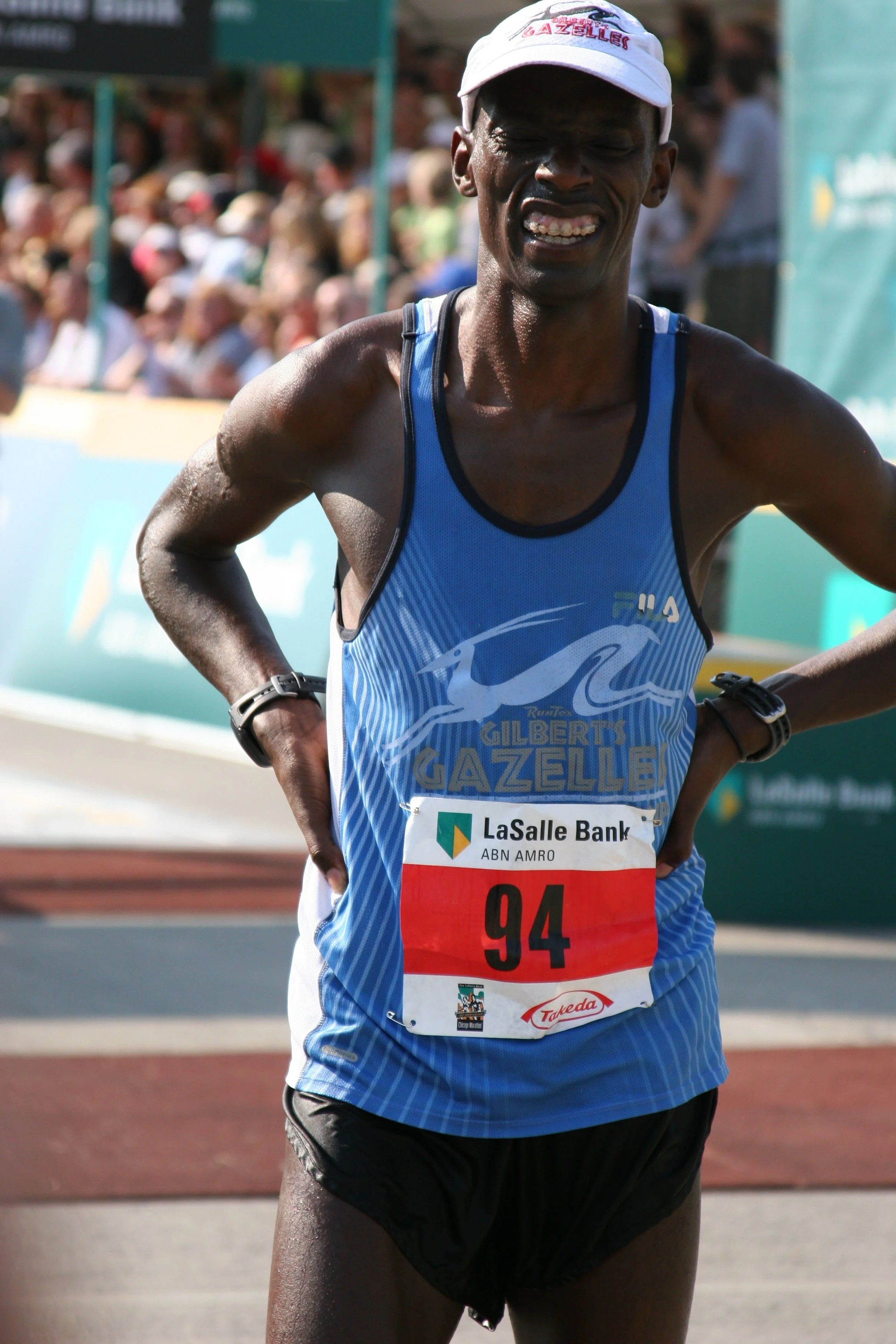 A male marathon runner wearing a blue Nike running tank top with the text Gilberts Gazelle, a white cap, black shorts, and a bib number 94, is finishing a race on a track with a crowd in the background.