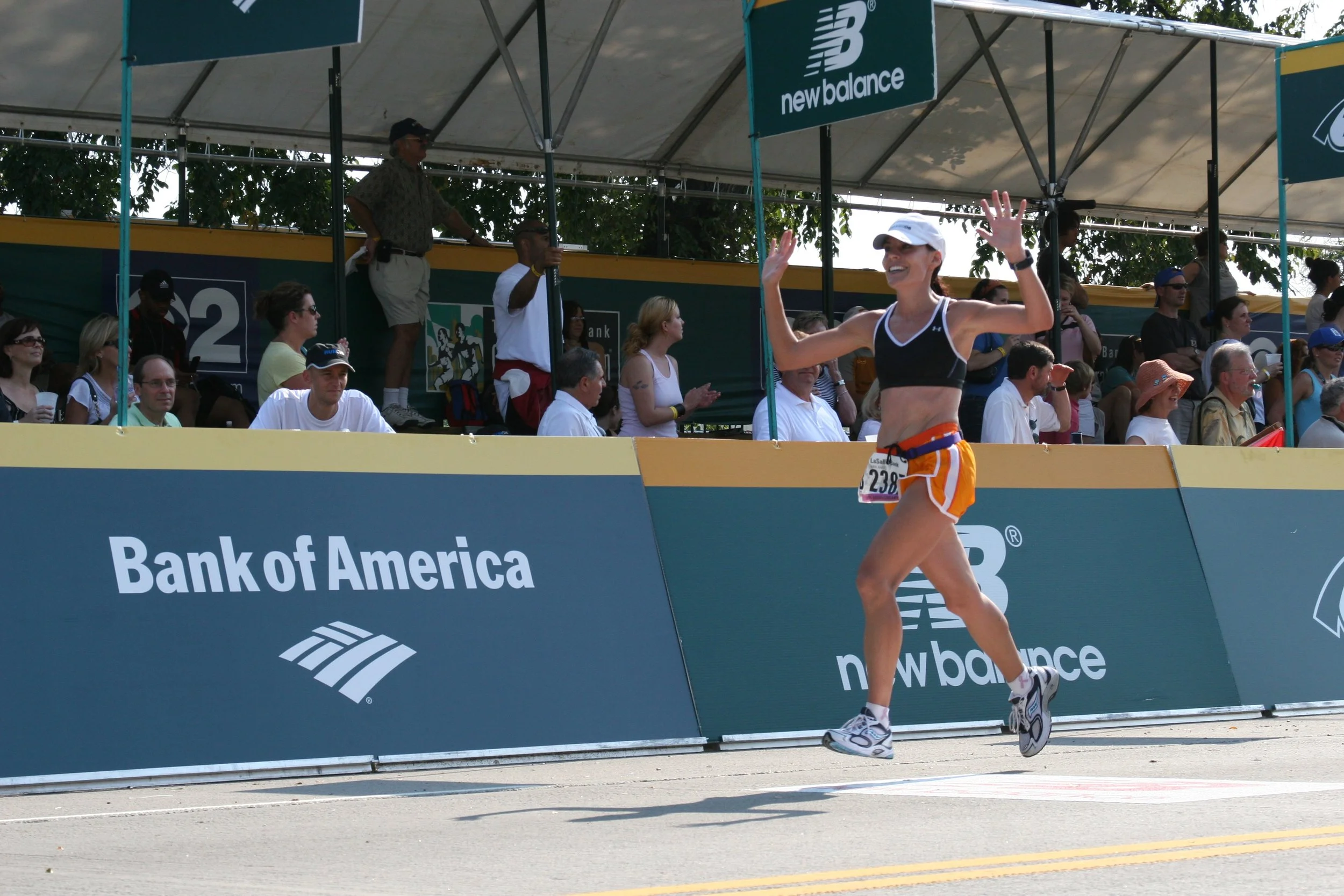 A woman runner wears a white cap, black sports bra, and orange shorts as she runs past the finish line at an event sponsored by Bank of America. Spectators and officials are seated and standing behind a barrier. The scene takes place outdoors under c