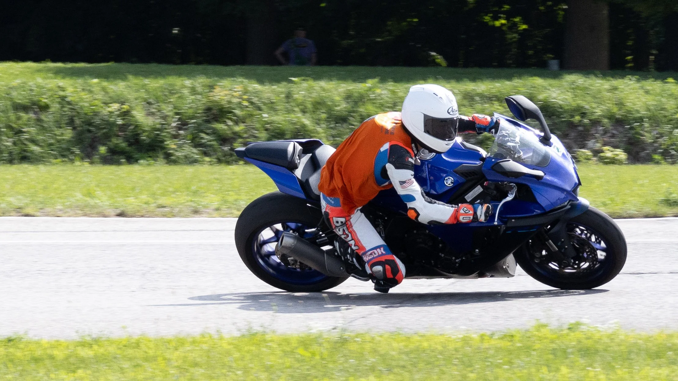 Motorcycle racer riding a blue bike on a road, wearing a white helmet and orange racing suit with black, white, and blue accents, while leaning into a turn.