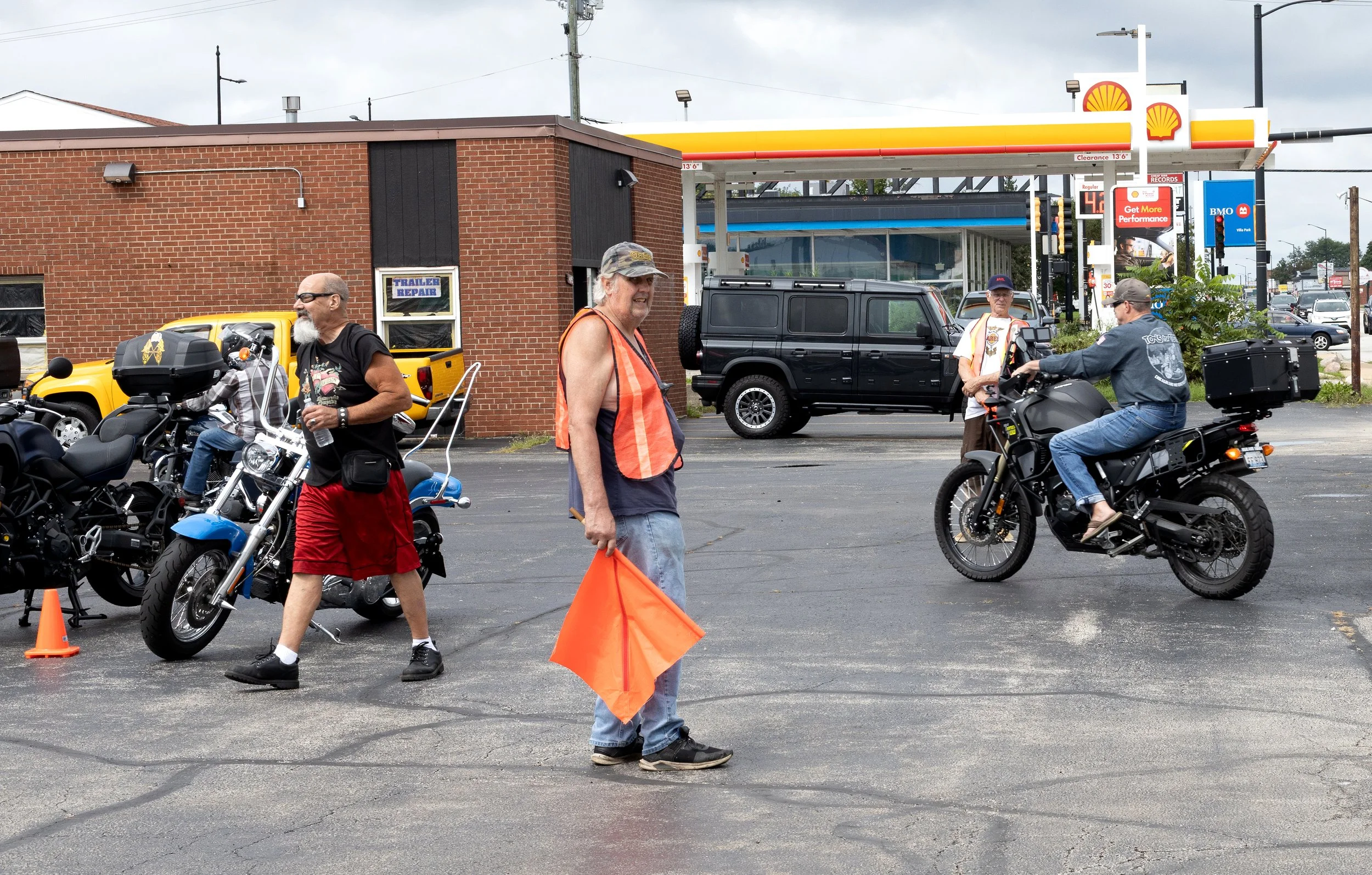 People gathered at a Shell gas station, some on motorcycles, with brick building and cars in the background.