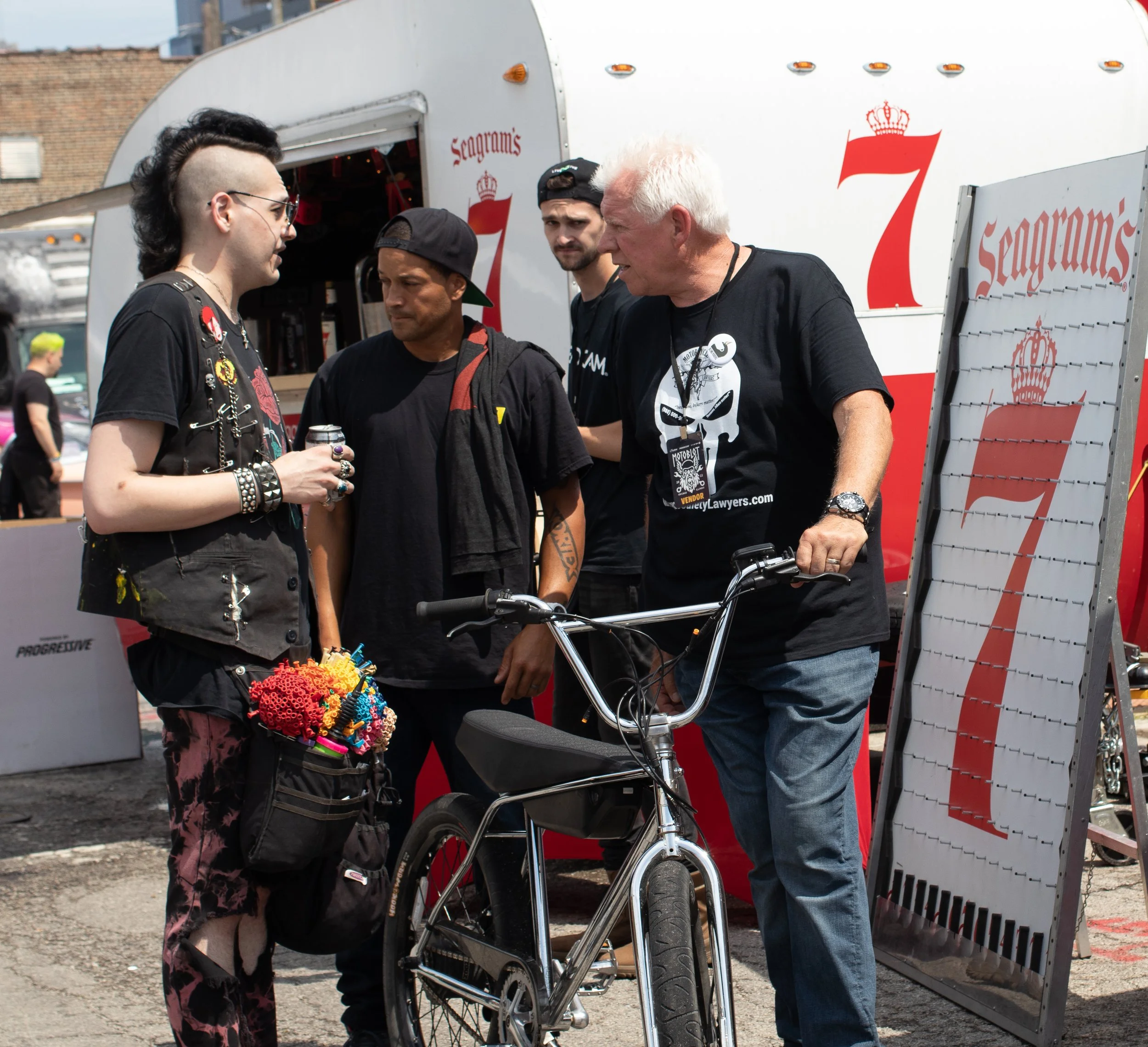 Four men are standing outdoors near a white trailer with red lettering and crown logo. One man with a mohawk hairstyle and sunglasses is holding a can, wearing a black T-shirt decorated with pins. The second man has a dark cap backwards, and the thir