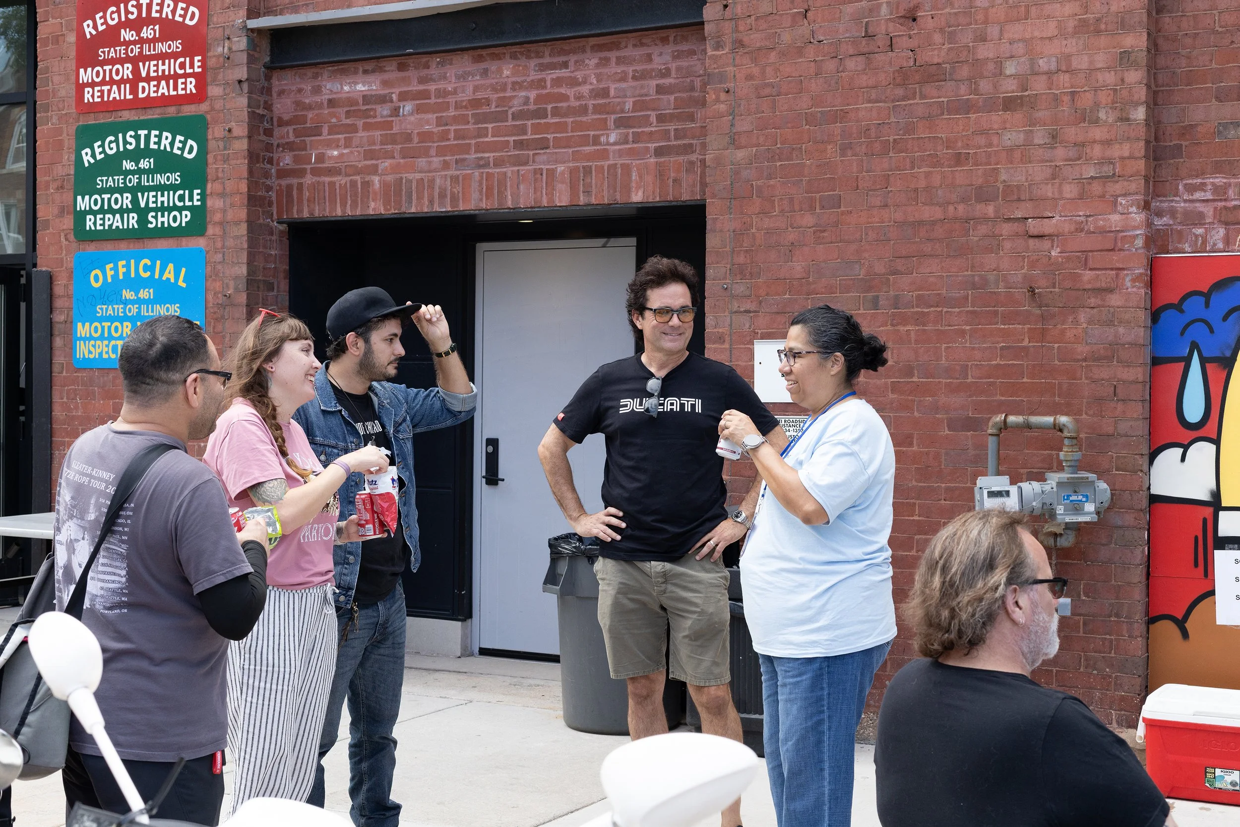 A group of people standing outside in front of a brick building, engaged in conversation. They are near signs indicating motor vehicle services and an illustration of a soda with a straw on a red vending machine.