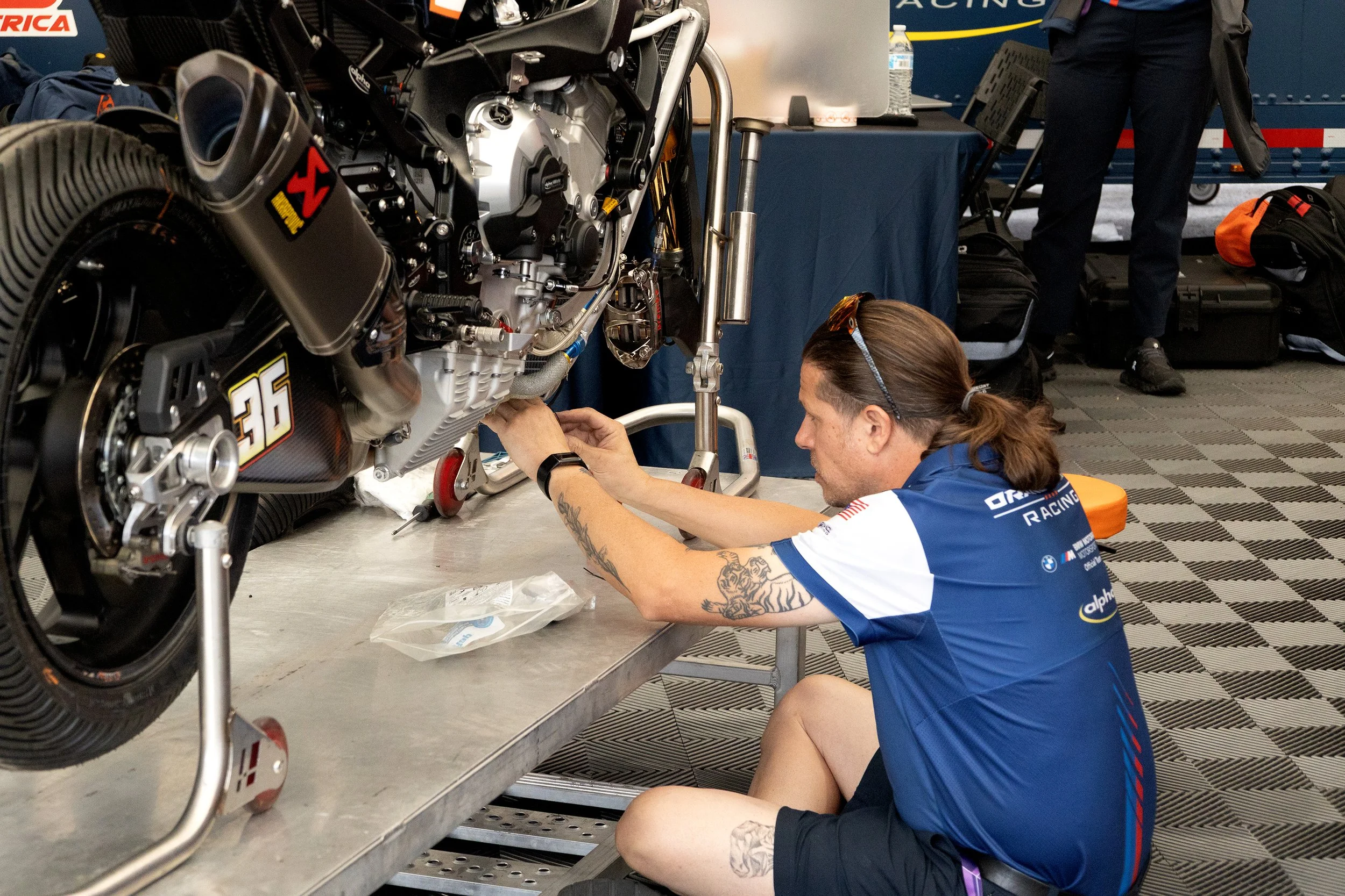 A person working on a motorcycle in a garage or workshop. The person has tattoos on their arms, is wearing a blue racing shirt and glasses on their head. The motorcycle is elevated on a stand, and there are various tools and equipment around.