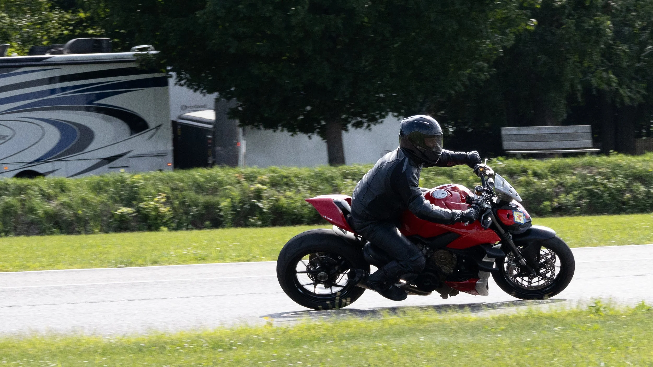 A person riding a red motorcycle on a paved road with green grass and bushes, and a dark-colored truck or RV and trees in the background.